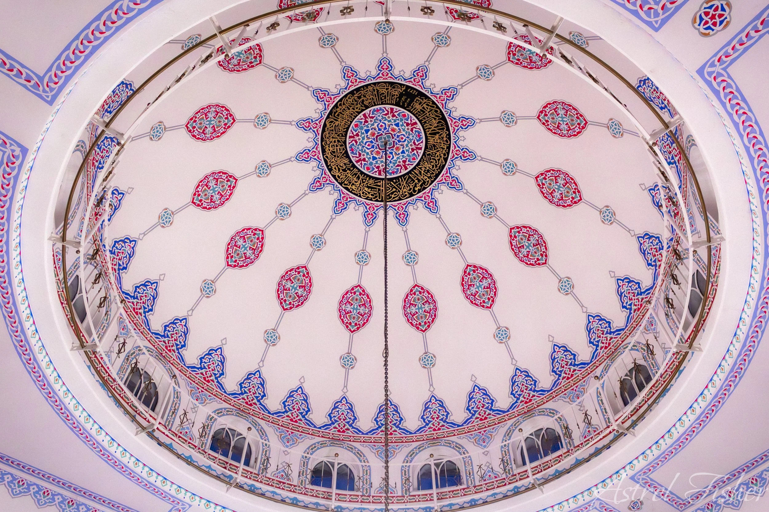 Decorative ceiling with intricate Islamic geometric patterns and calligraphy in gold, red, blue, and white colors around a central black circular design.