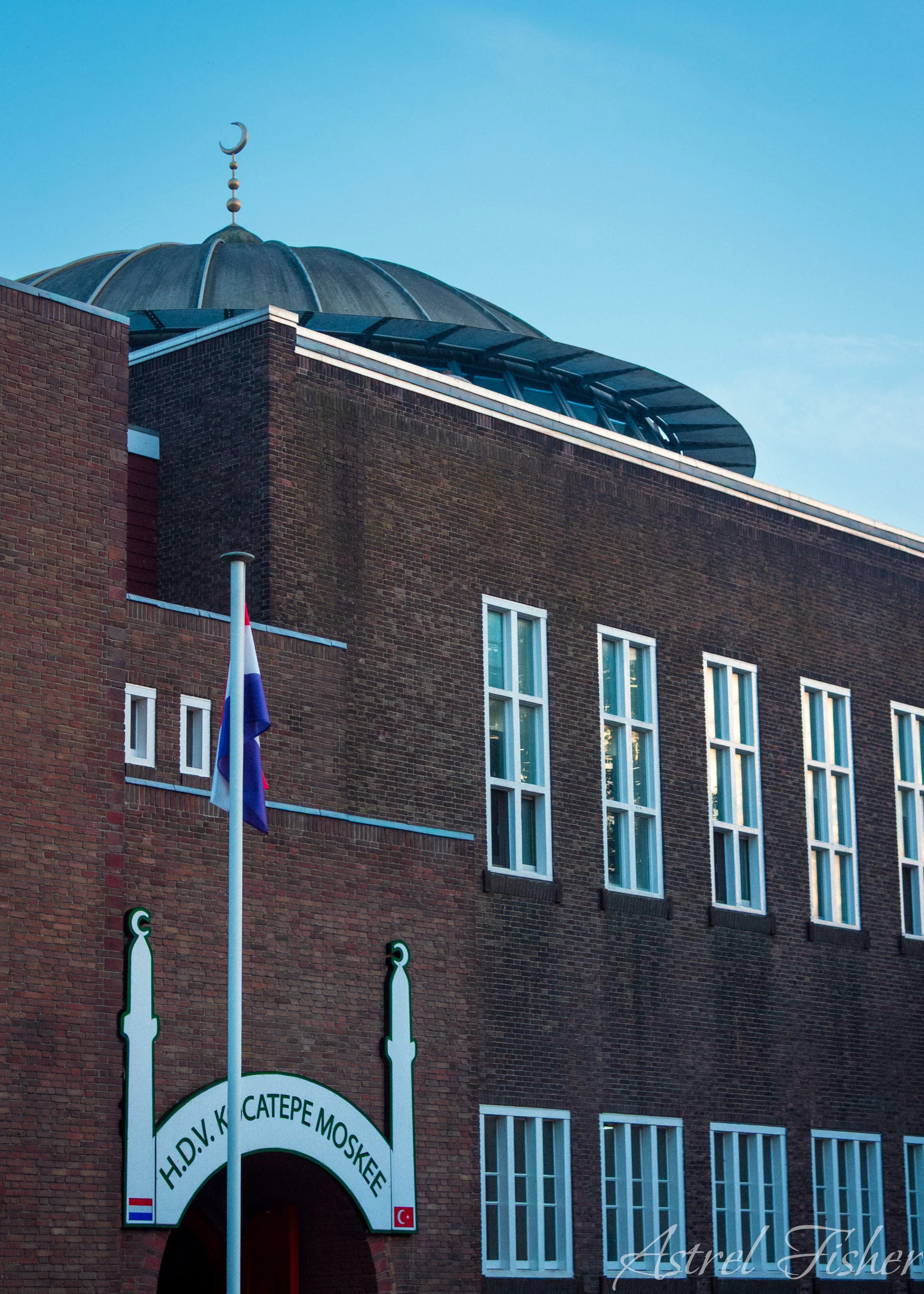 A brick building with a minaret topped with a crescent moon symbol. A sign reading 'H.D.V. K. CATER E MO' is visible below the archway entrance. A flagpole with a flag is in front of the building. The windows are rectangular and have vertical panes.