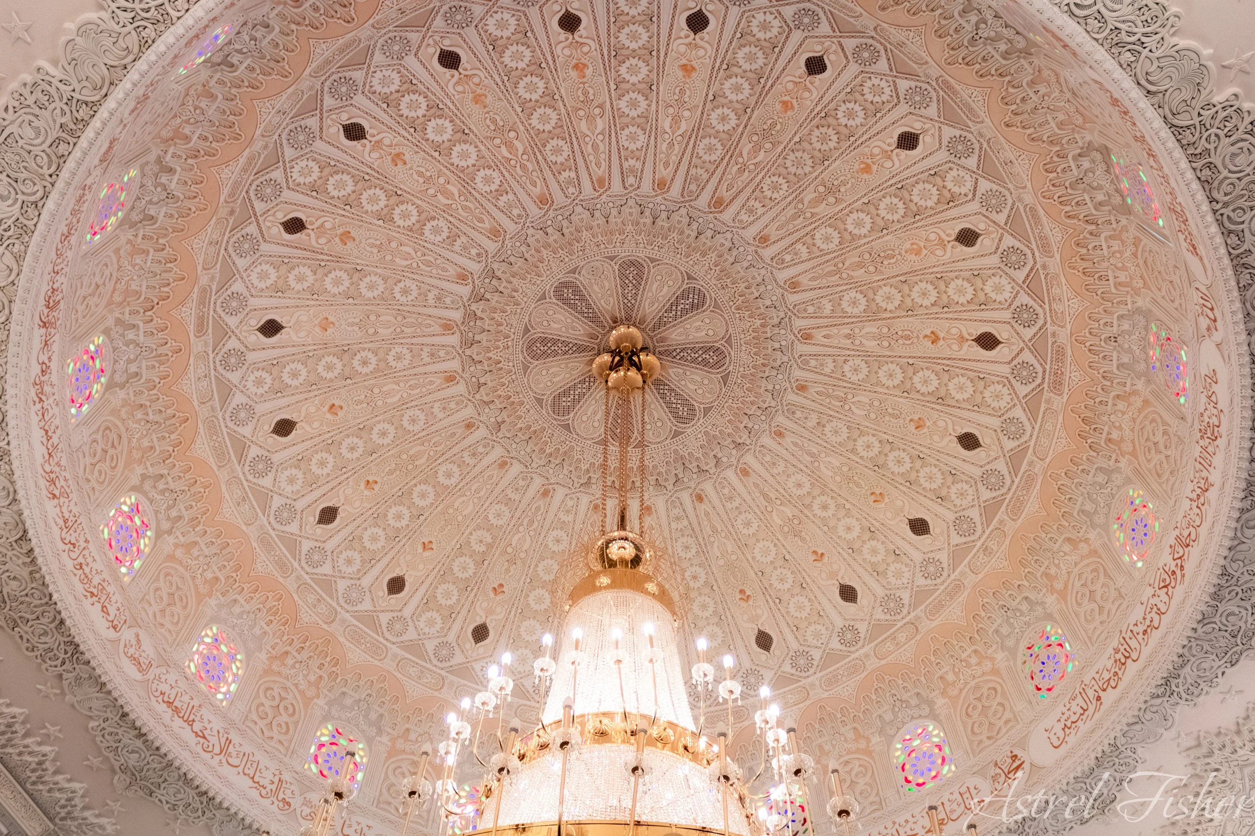 An ornate, decorative ceiling with intricate patterns and Arabic calligraphy. A chandelier hangs from the center, illuminating the space with warm light. The ceiling design includes floral and geometric motifs with pastel colors and gold accents.