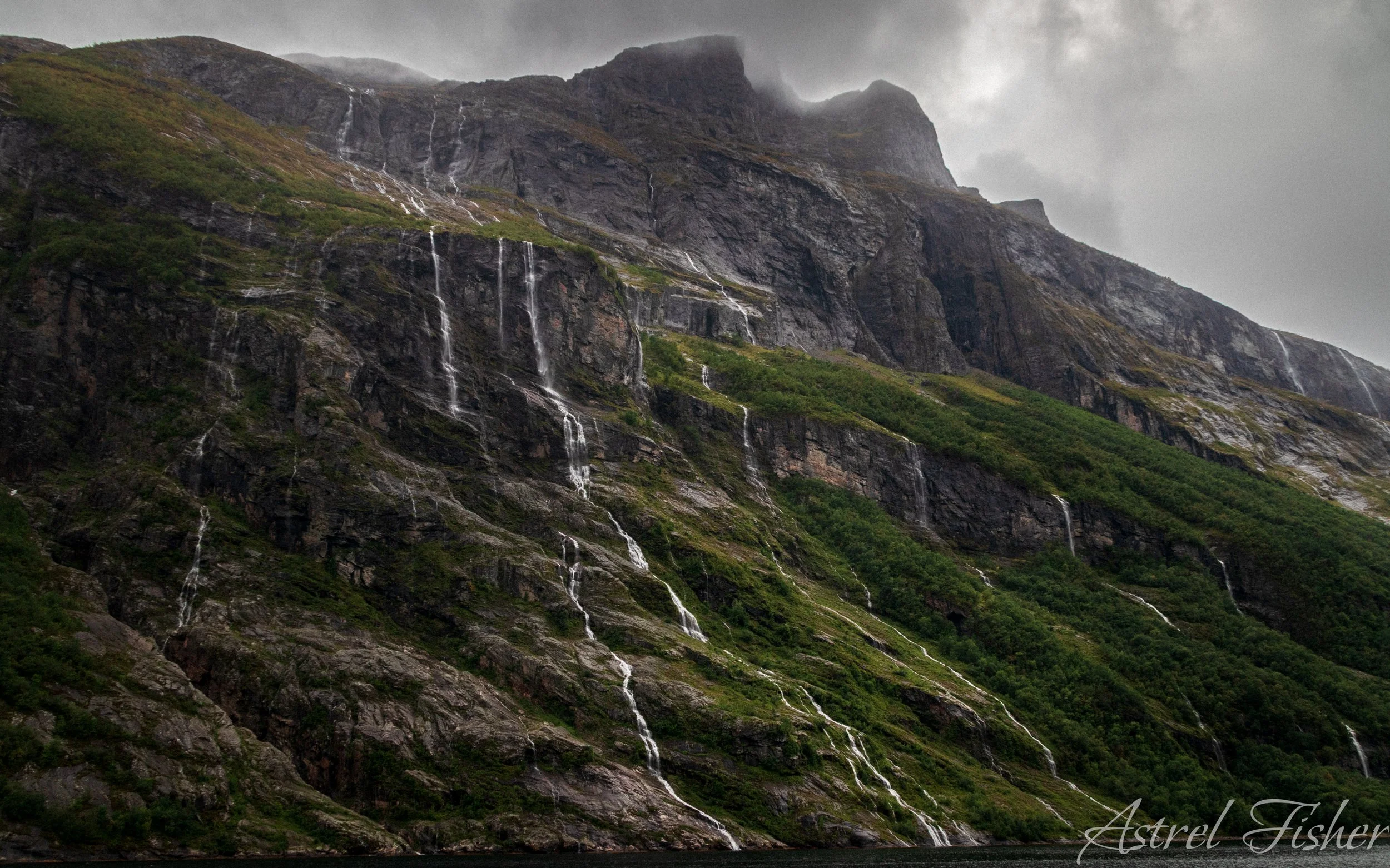 A large mountain with multiple waterfalls cascading down its rocky slopes, partially covered by clouds, with some greenery at the base.