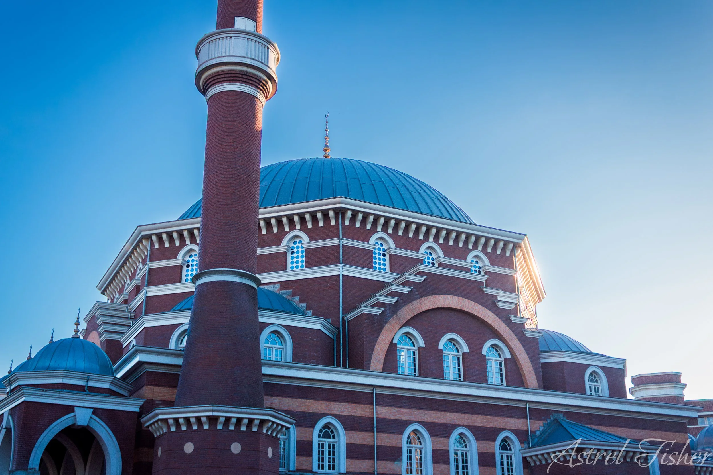 A large red brick building with multiple blue domes and a tall minaret against a clear blue sky.