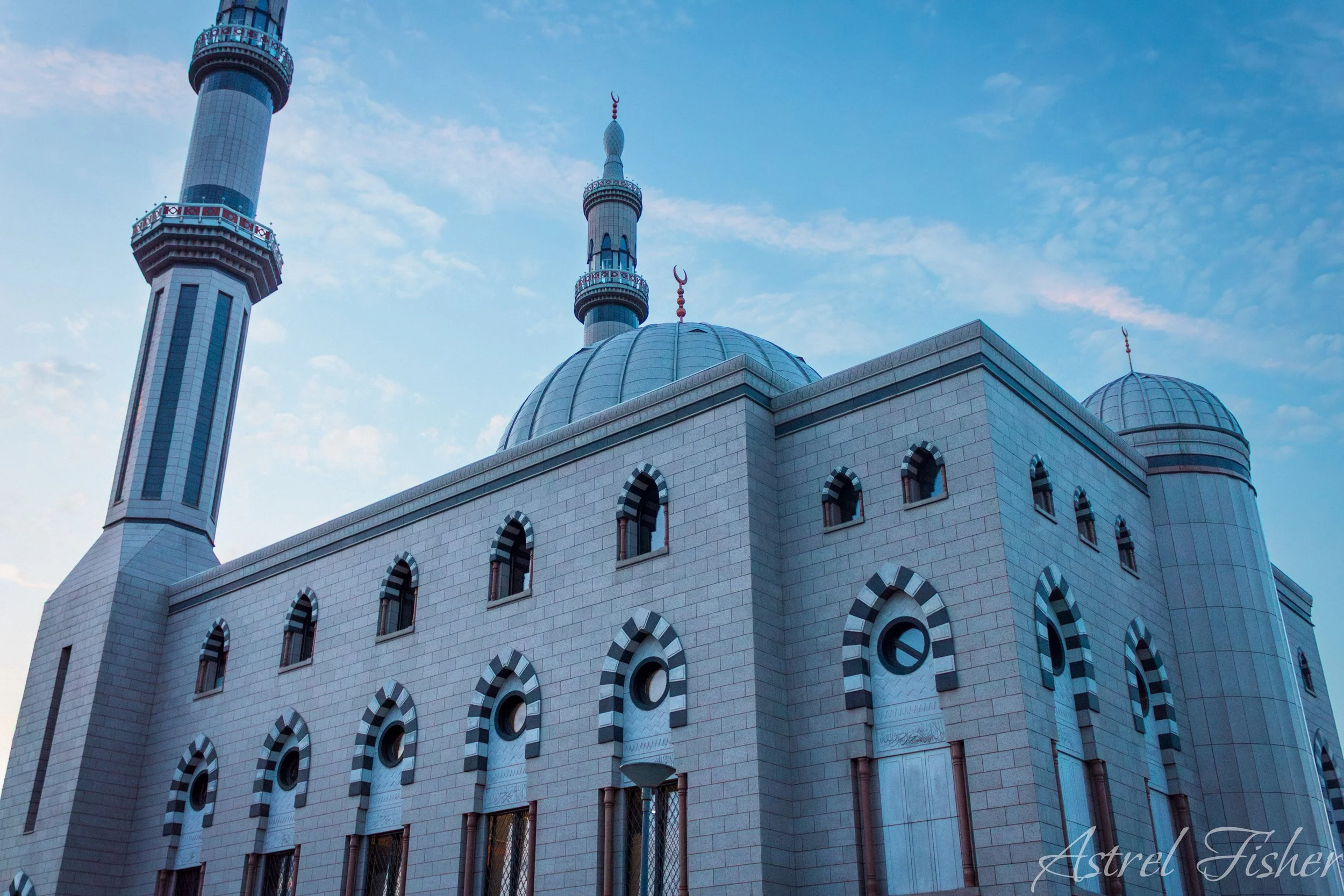 A mosque with a large dome, arched windows, and two minarets set against a blue sky with some clouds.