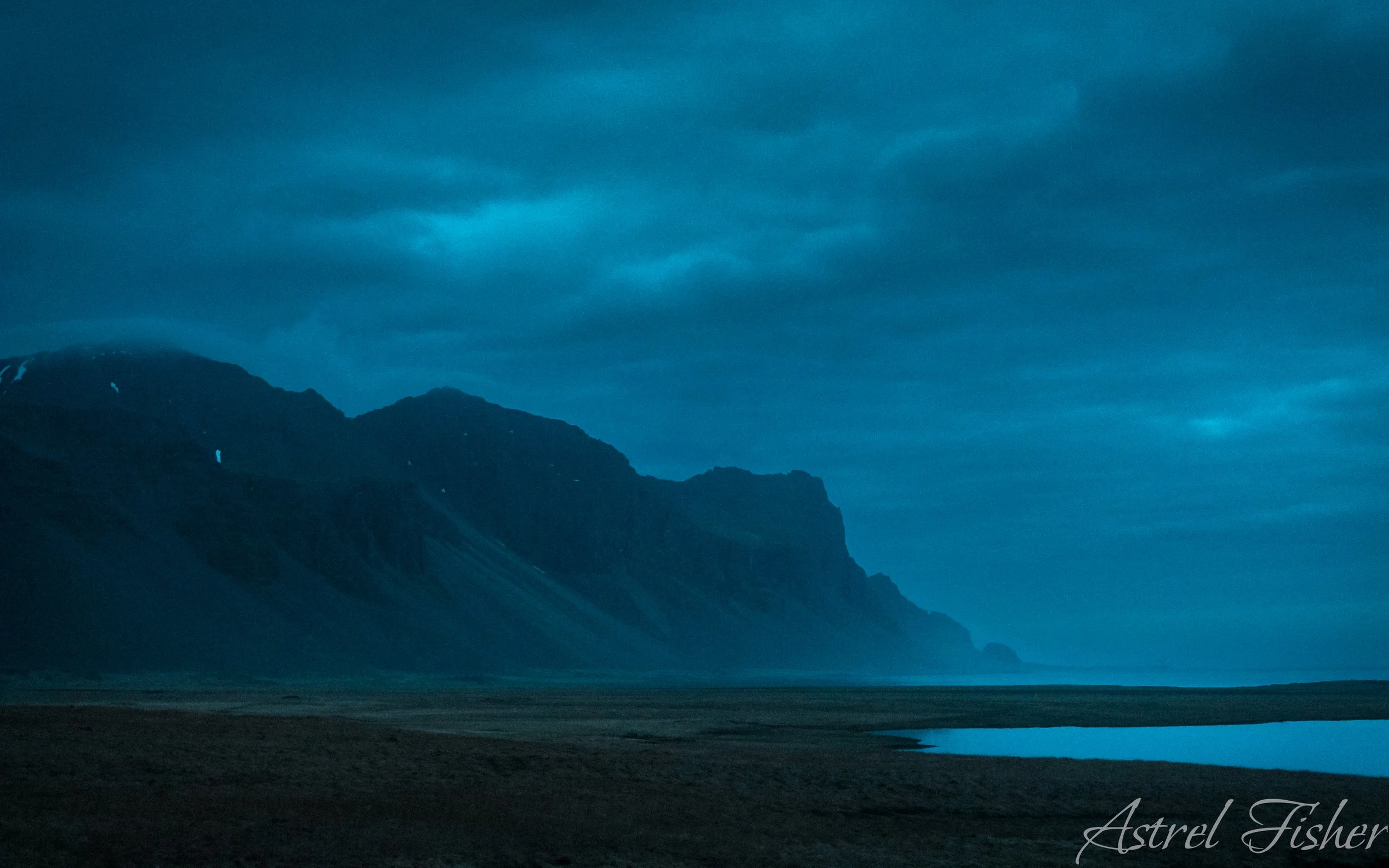 Mountain range under a cloudy, blue-toned sky with a body of water and flat land in the foreground.