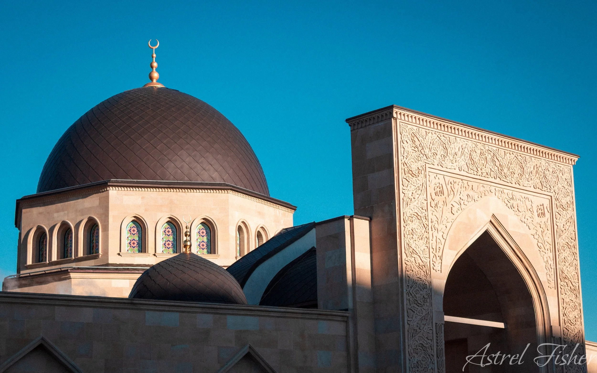 Close-up of architectural features of a mosque, including domes, windows with stained glass, and intricate carvings on the archway, under a clear blue sky.