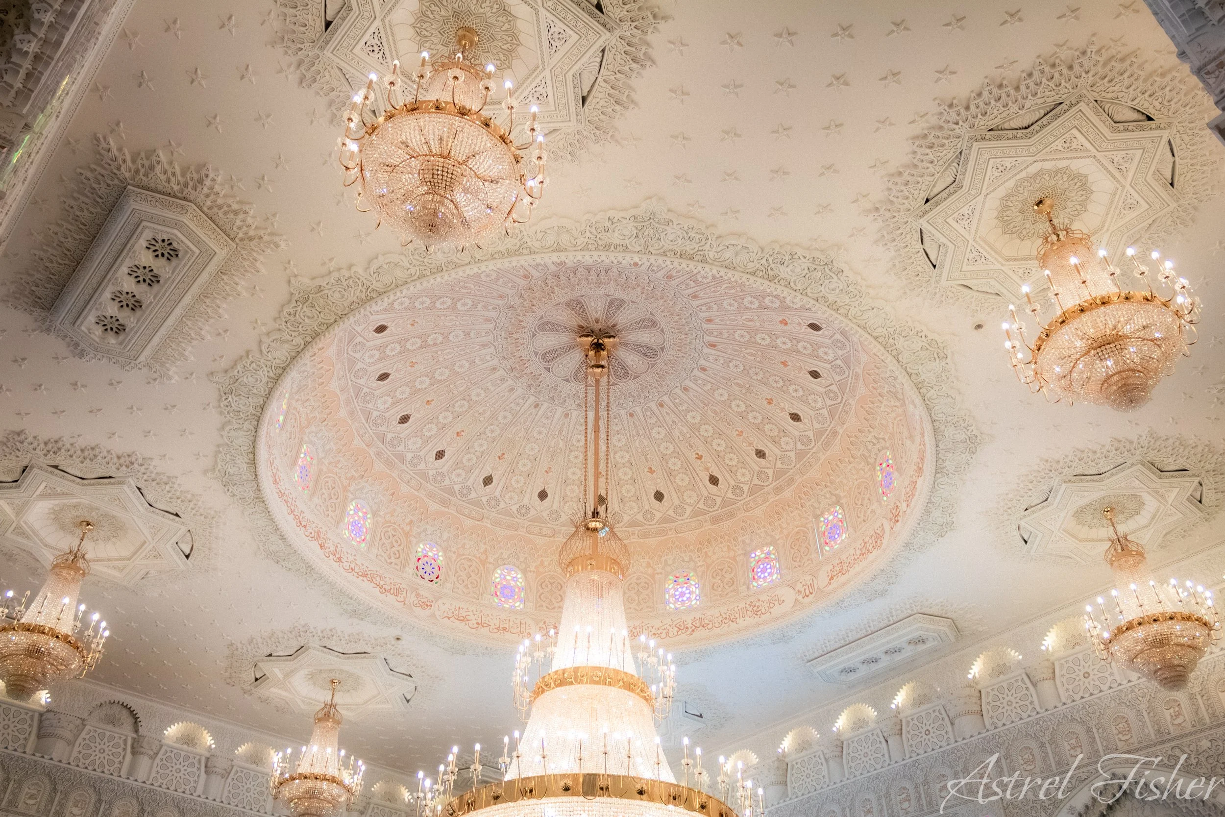 Intricate white ceiling with floral and star patterns, large ornate chandeliers, and stained glass windows with Arabic calligraphy.
