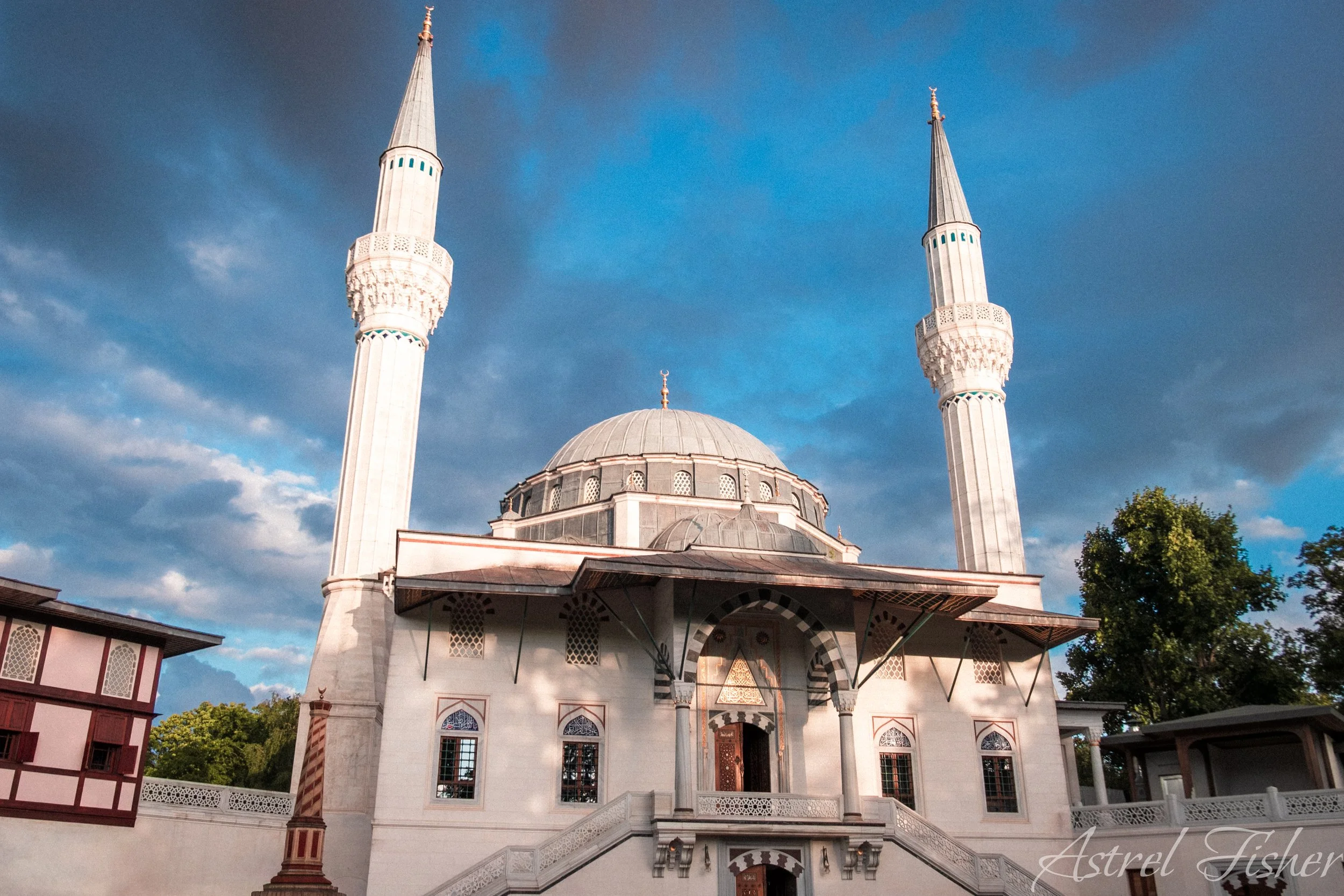 A mosque with a large central dome and two tall minarets against a partly cloudy sky, with trees surrounding the building.