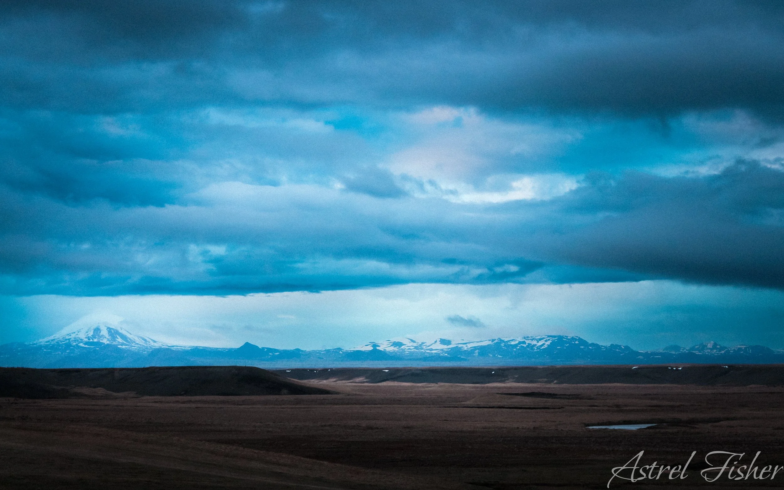 Scenic landscape with snow-capped mountains in the distance, dark cloudy sky, and flat plains in the foreground.