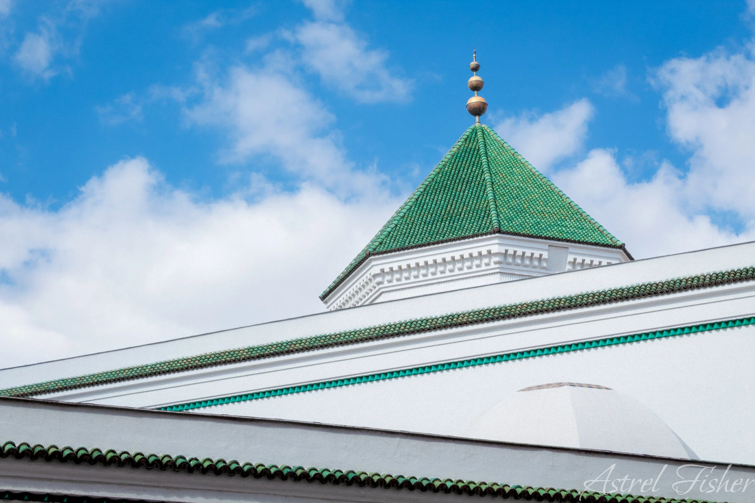 Close-up of a building with a green tiled roof and white walls, topped with a decorative spire against a blue sky with scattered clouds.
