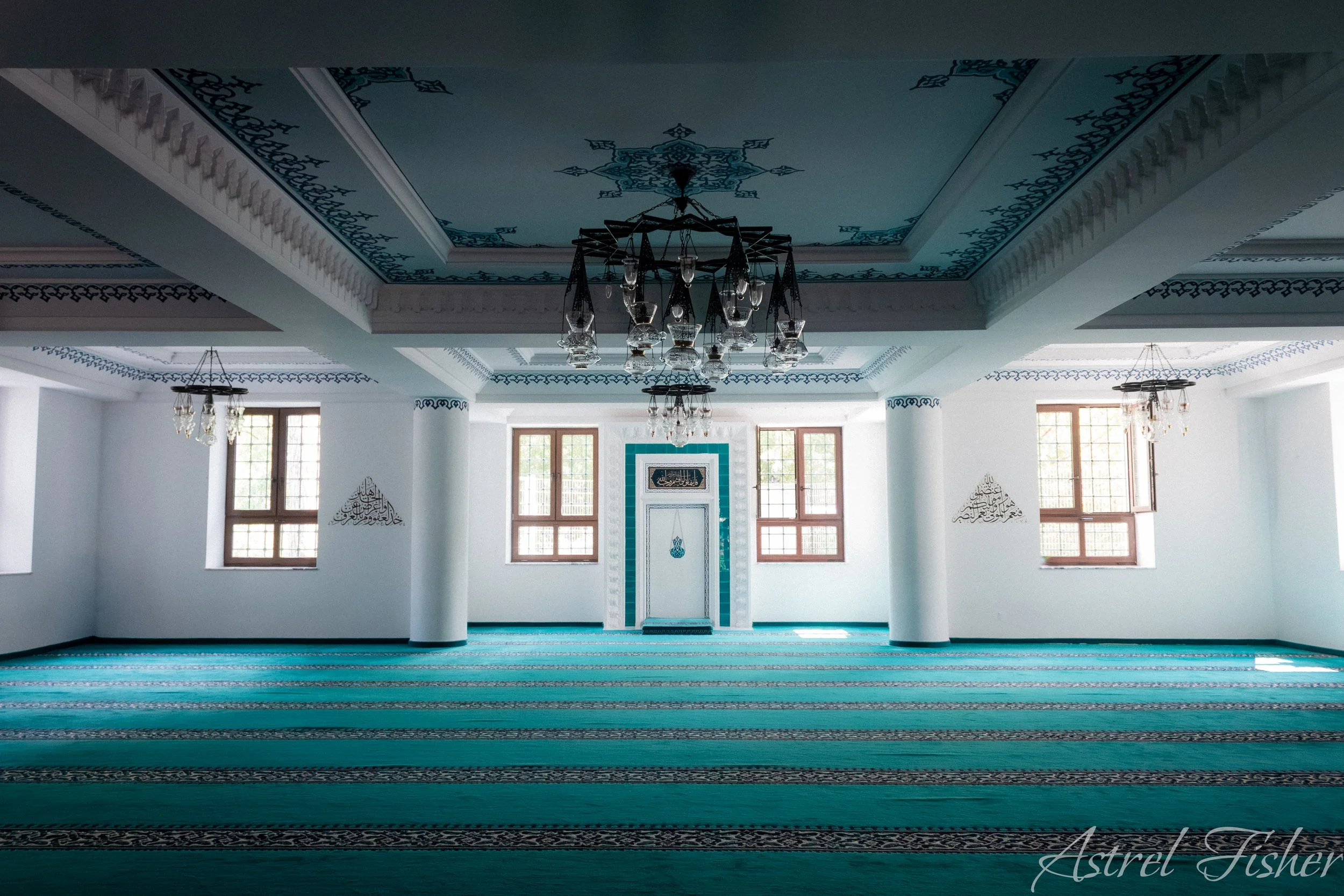 Interior of a mosque with a turquoise prayer rug, white columns, windows, chandeliers, and Islamic calligraphy on the walls.