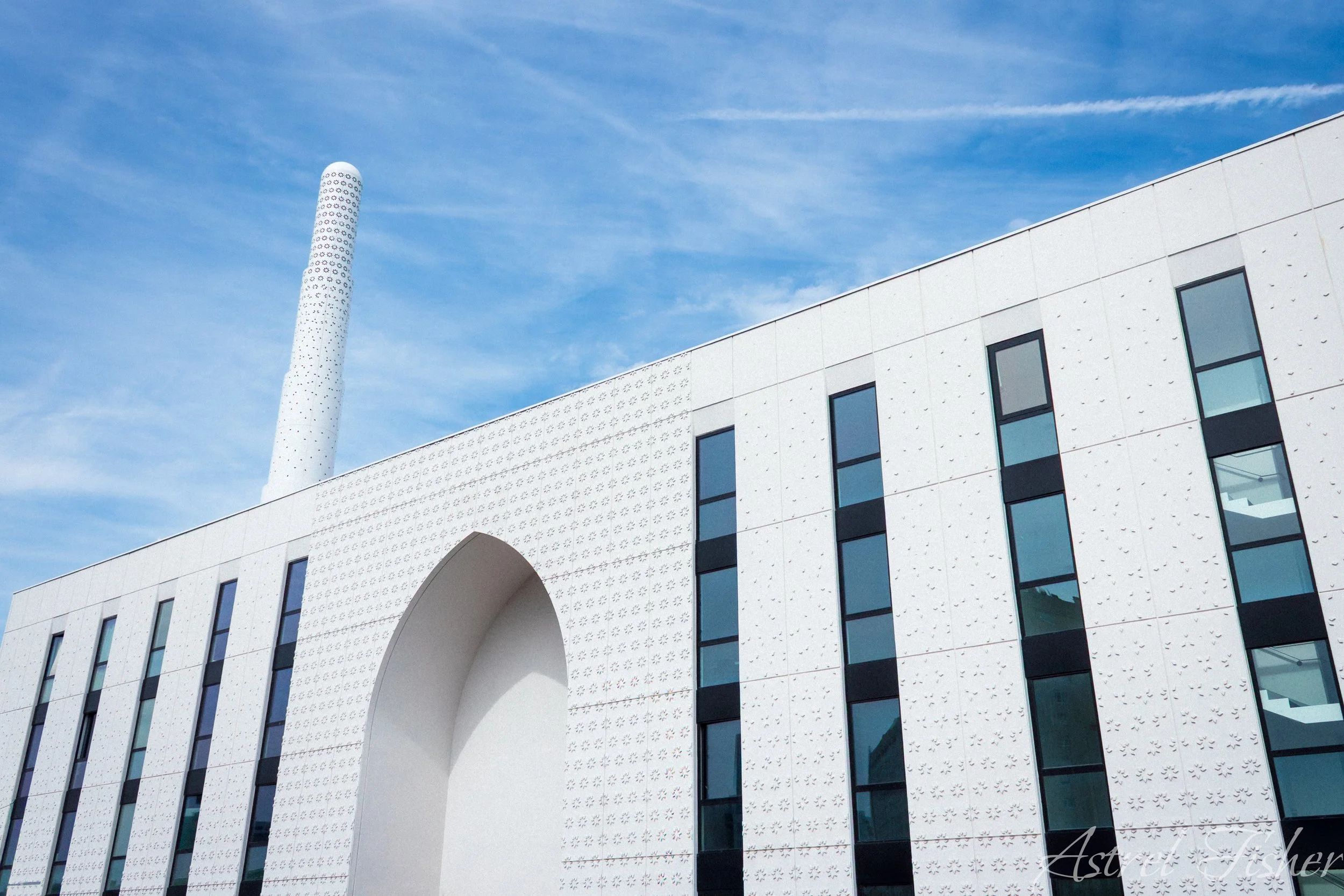 Modern white building with black window frames and a decorative archway, with a tall patterned chimney against a blue sky with wispy clouds.