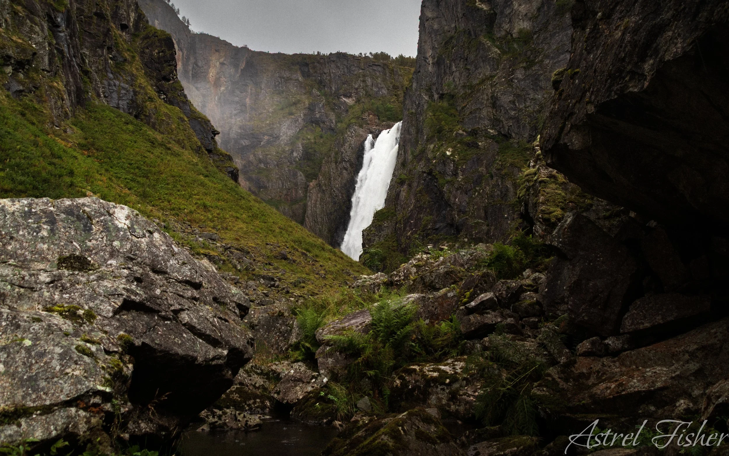 A waterfall flowing down a steep rocky canyon with lush green moss and plants on the rocks, overcast sky.