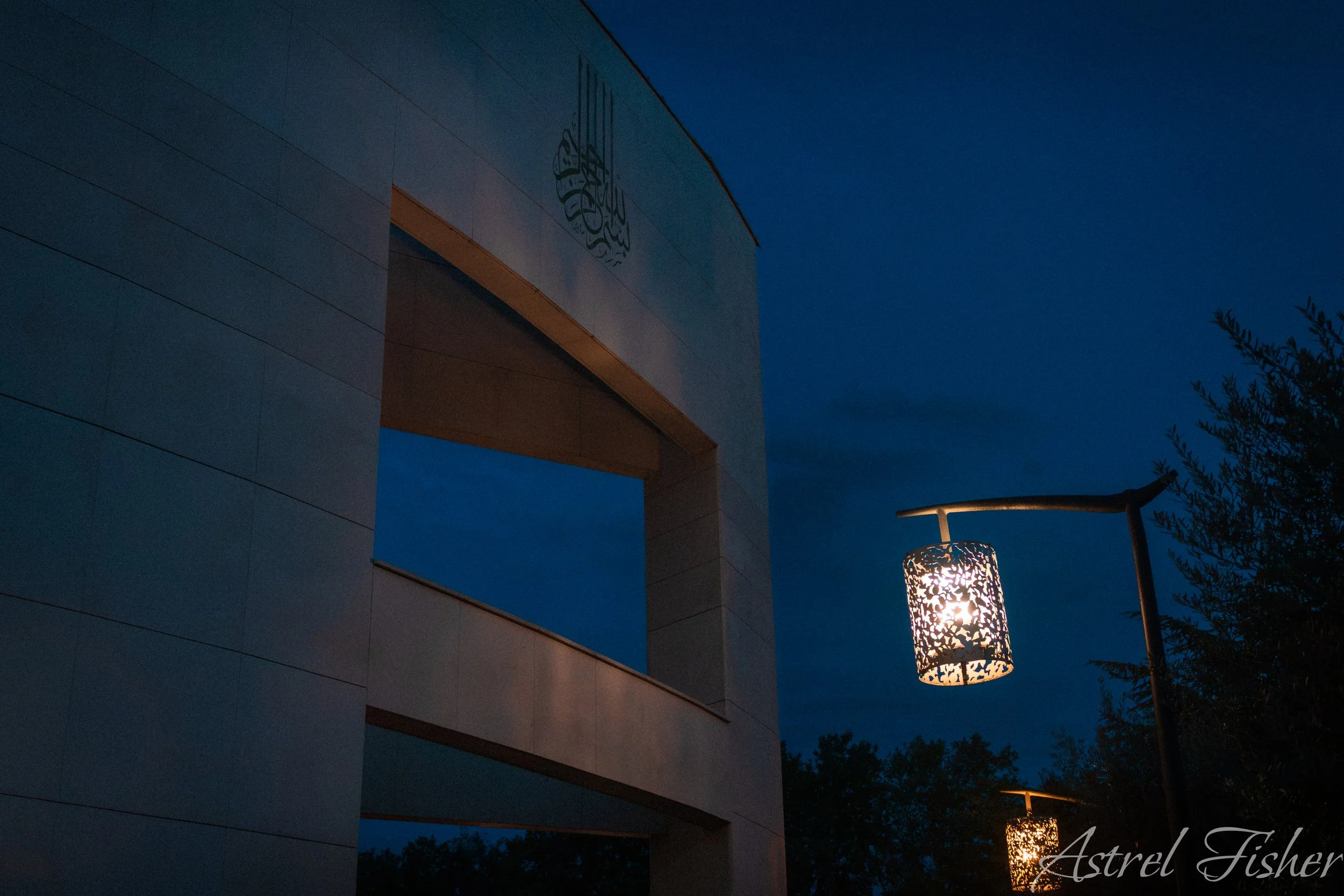 A modern building with curved white exterior walls, a decorative lamp hanging from a pole, and trees in the background at dusk.