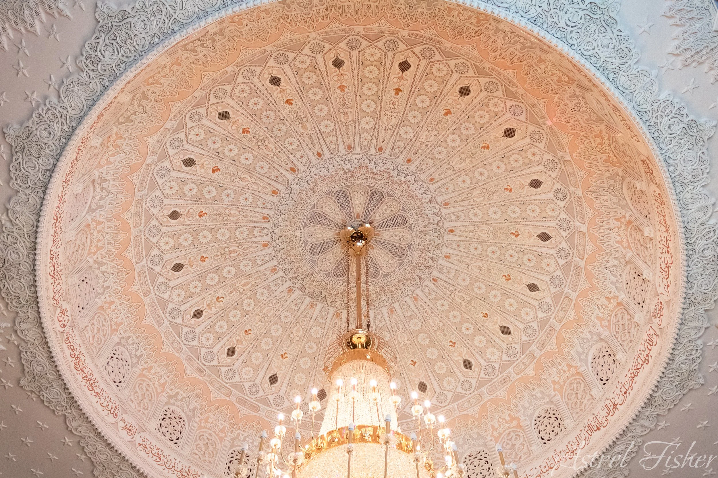 Interior view of an ornate, decorated ceiling with intricate white and peach designs, and chandeliers hanging below.