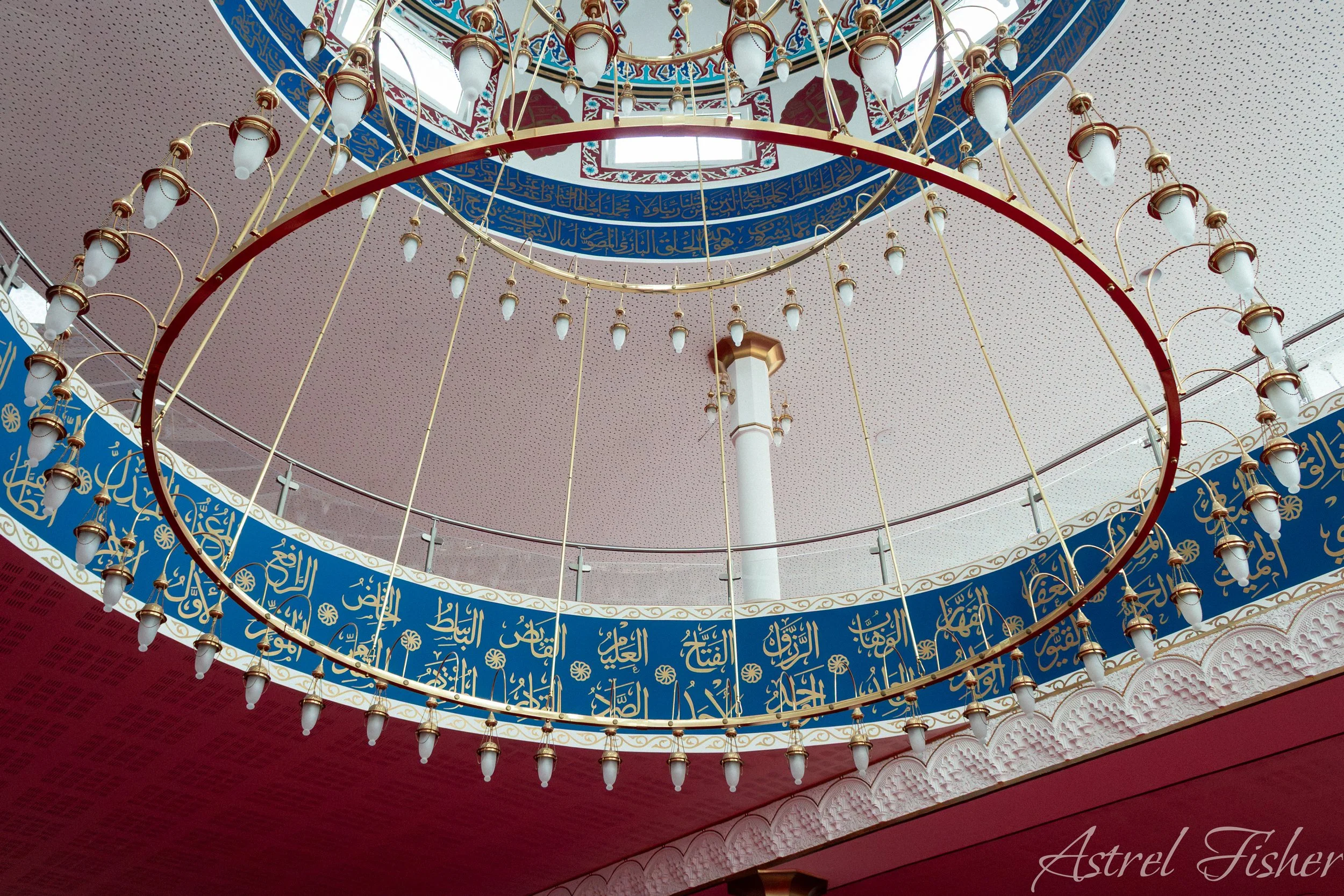 Interior view of a mosque ceiling featuring circular chandeliers, ornate Arabic calligraphy in blue and gold, and a central dome with windows for natural light.