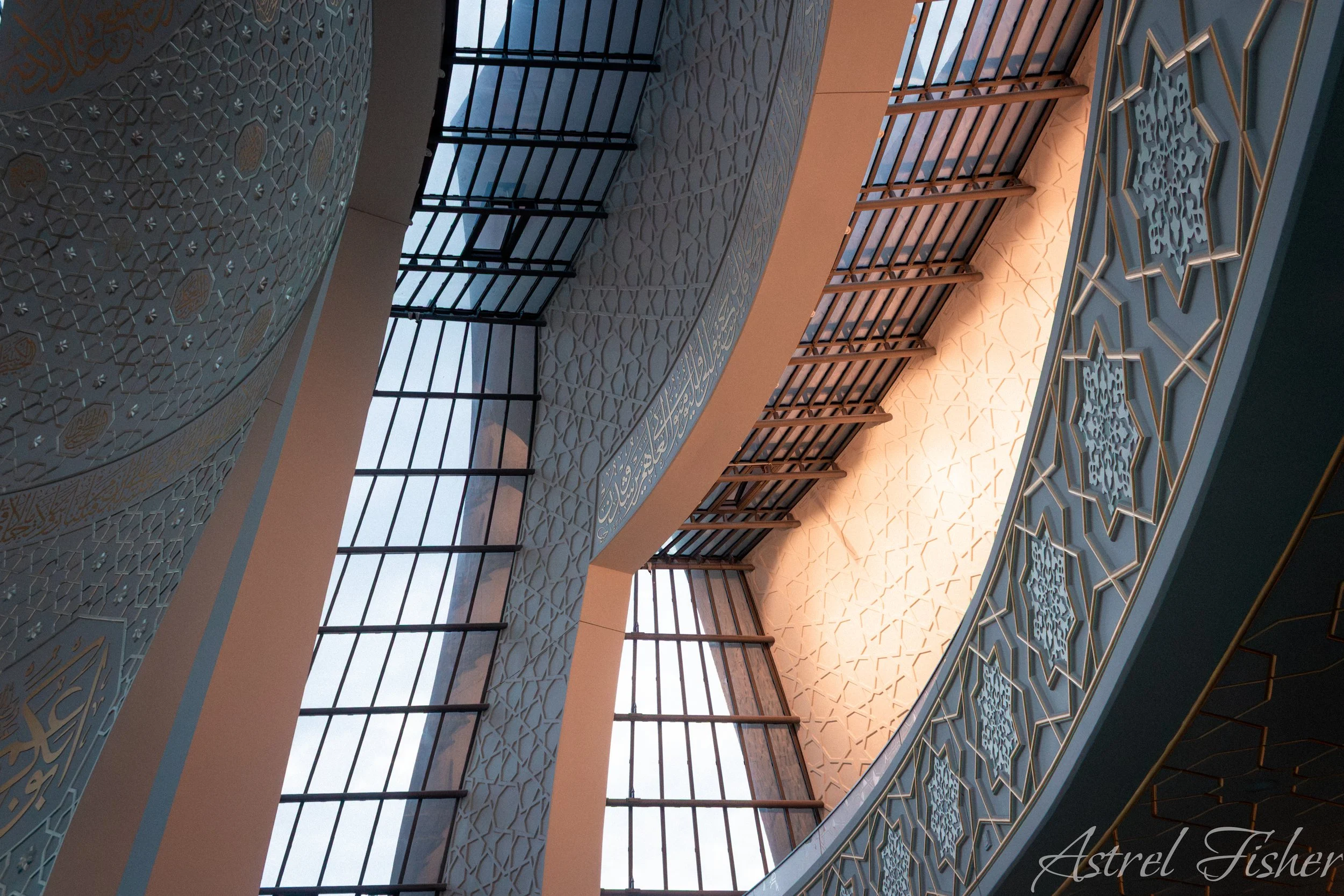 Interior view of a building with Islamic architectural design, featuring intricate geometric patterns and decorative elements on the ceiling and walls, along with large windows and wooden railing.