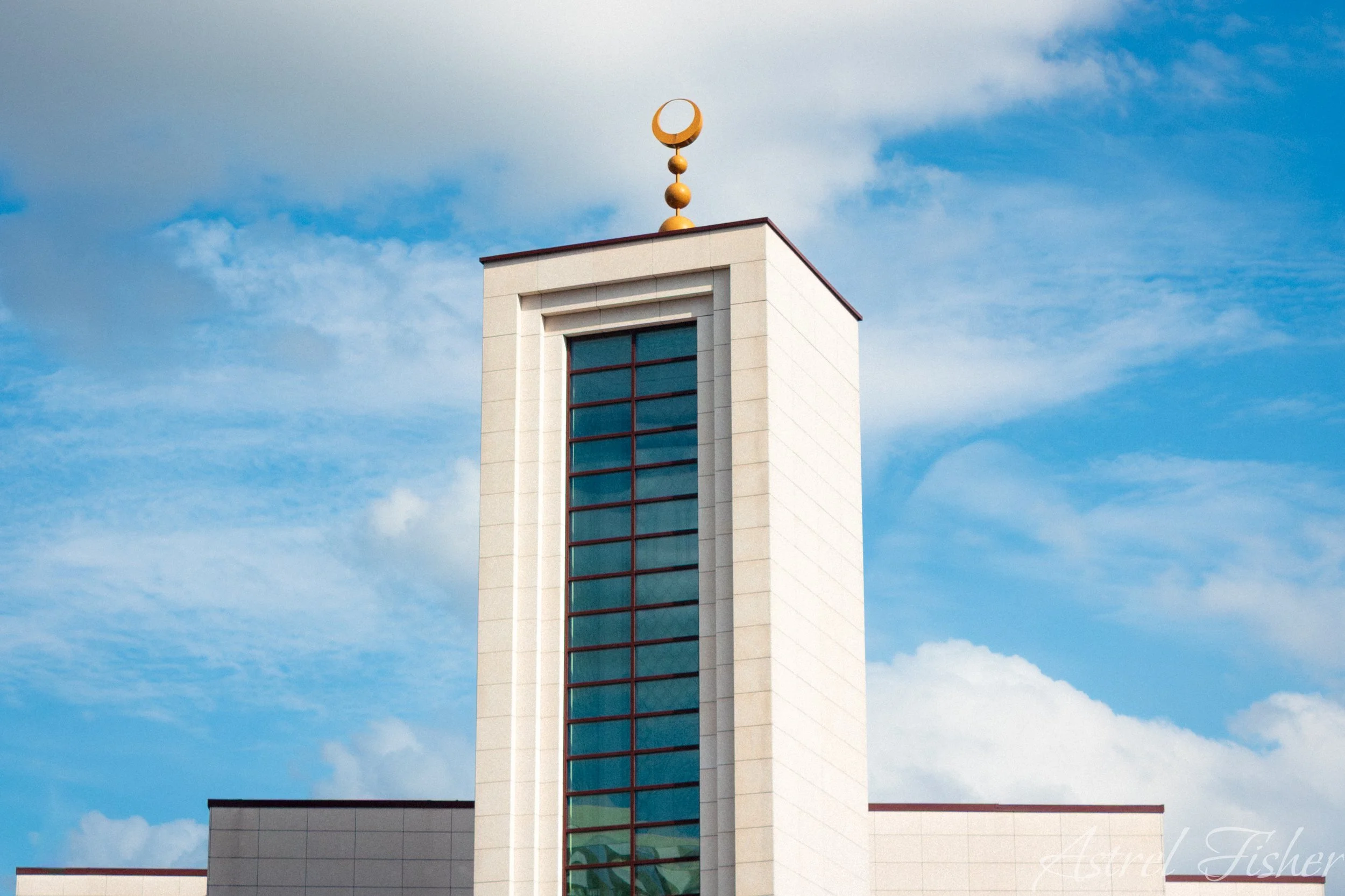 A modern white building with a large vertical glass window and a decorative golden element on top, set against a blue sky with scattered white clouds.