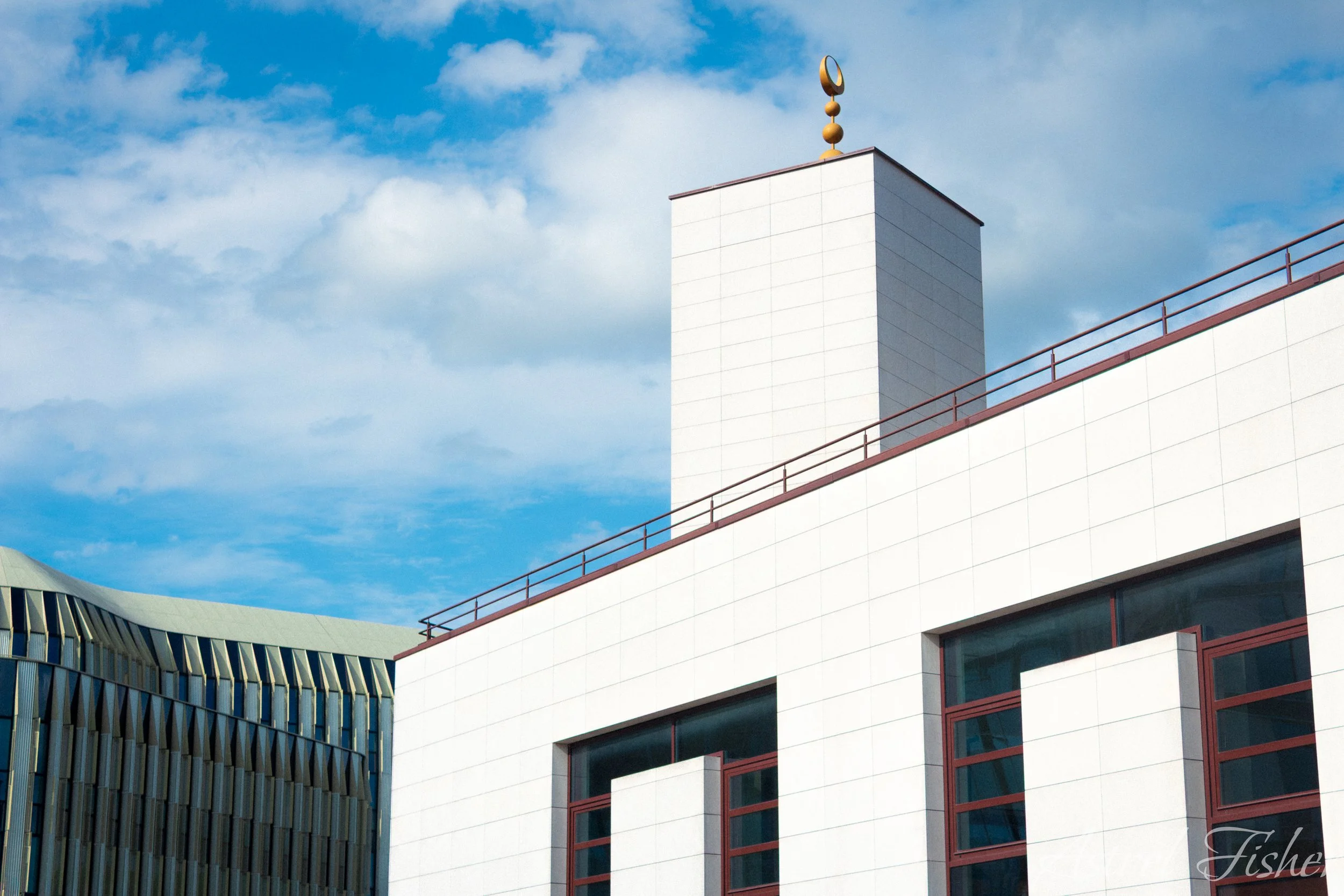 Close-up of modern white building with red-brown railings and glass windows under a blue sky with scattered clouds.