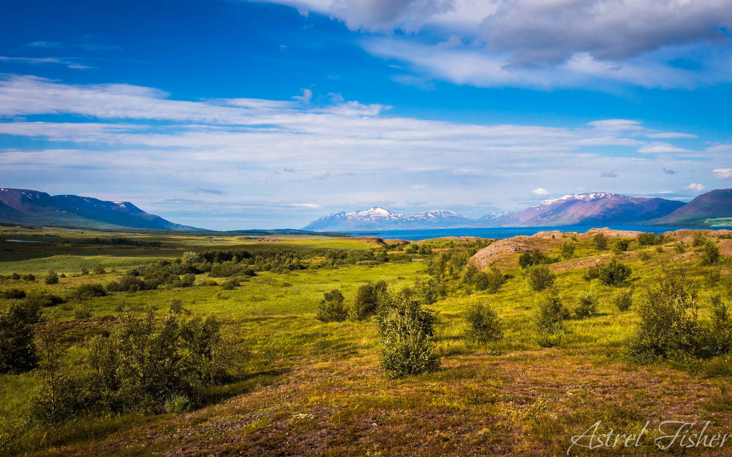 Scenic landscape with green fields in foreground, distant mountains with snow patches, calm blue lake between mountains, and partly cloudy sky.