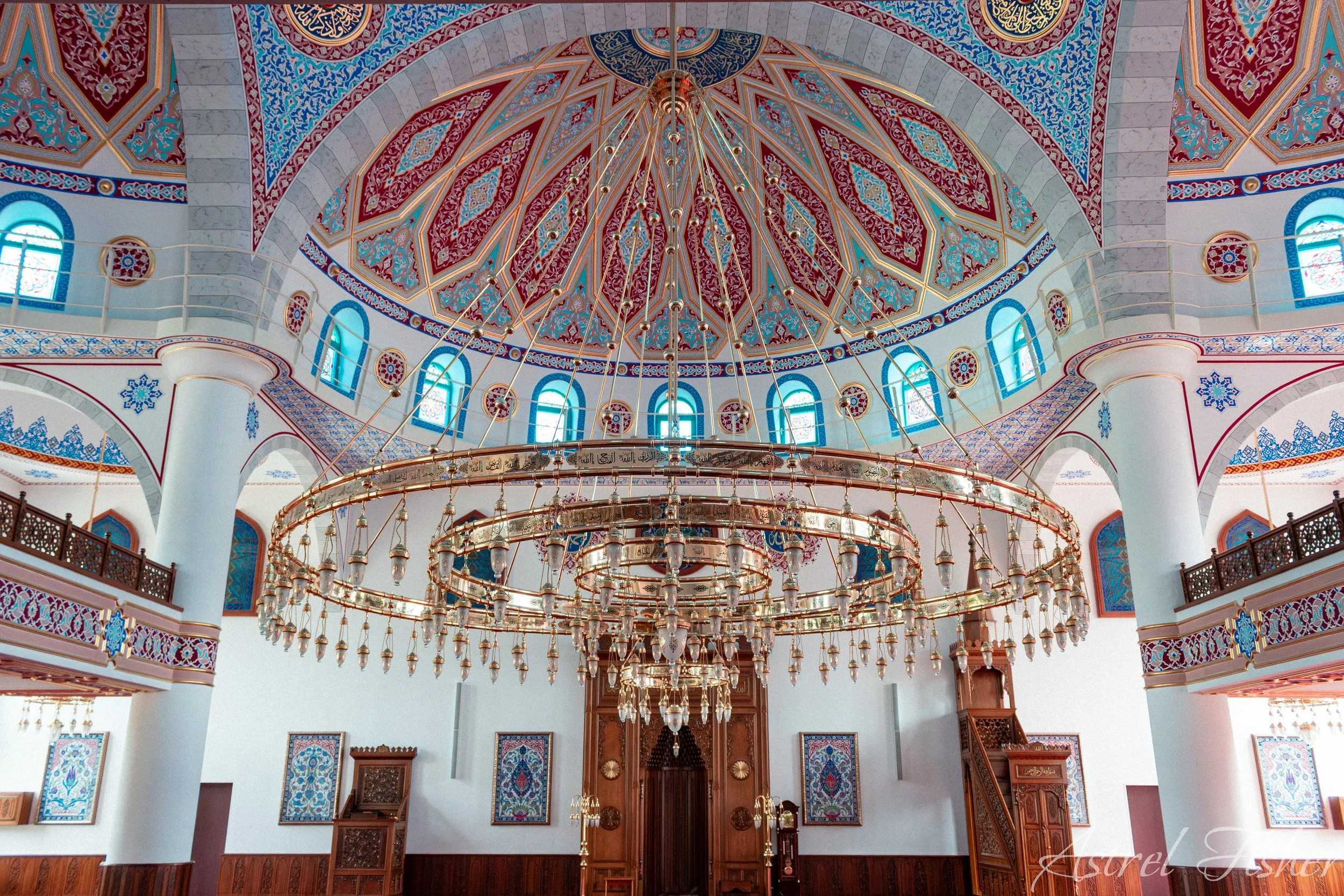 Inside a mosque with a large, ornate chandelier hanging from a decorative, intricately patterned domed ceiling with red, blue, and gold designs. White columns support the structure, and there are windows with colorful stained glass around the perimet