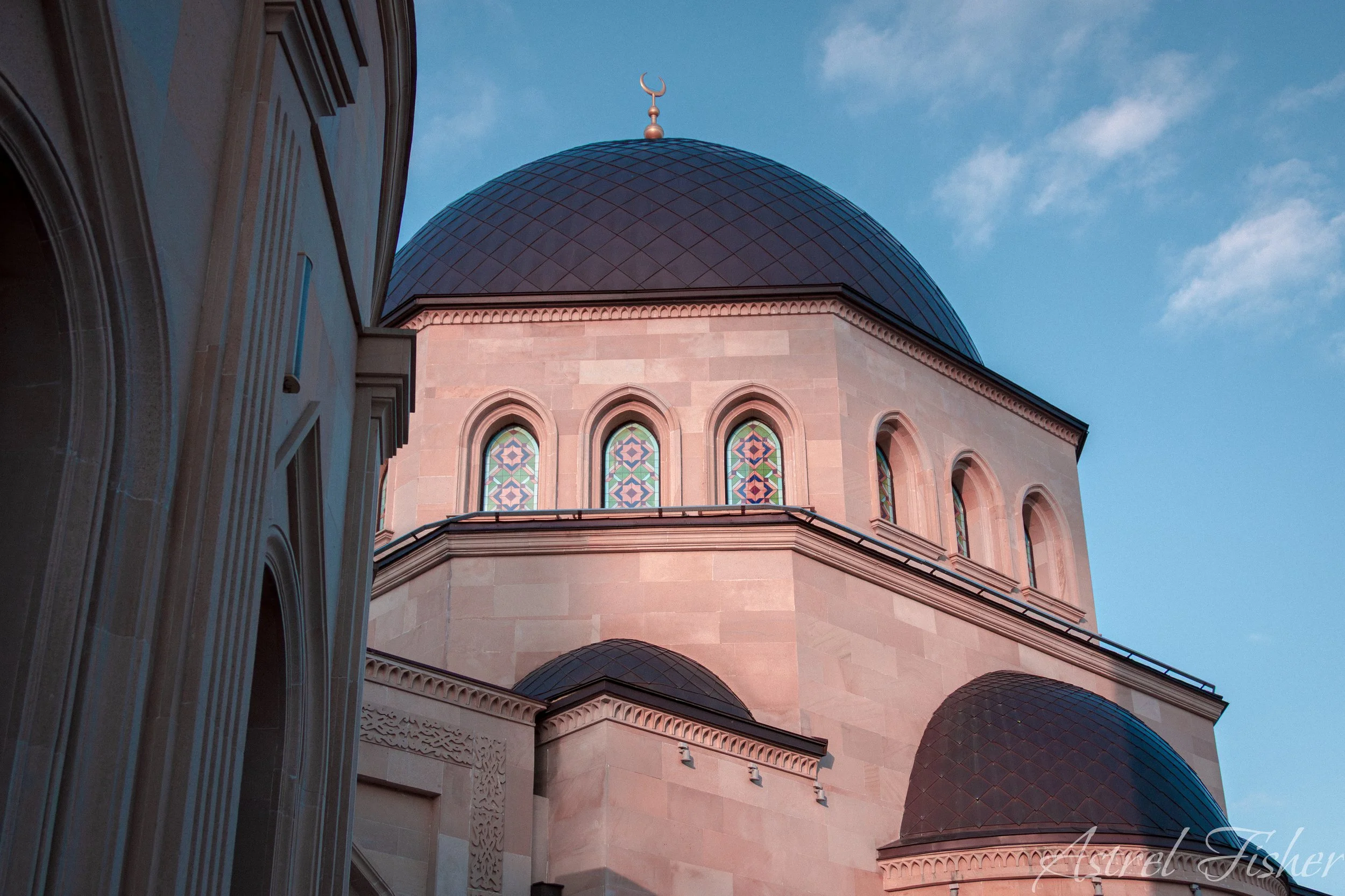 The photo shows a close-up view of a mosque or Islamic temple with stained glass windows and a dome topped with a crescent moon symbol, against a blue sky with some clouds.