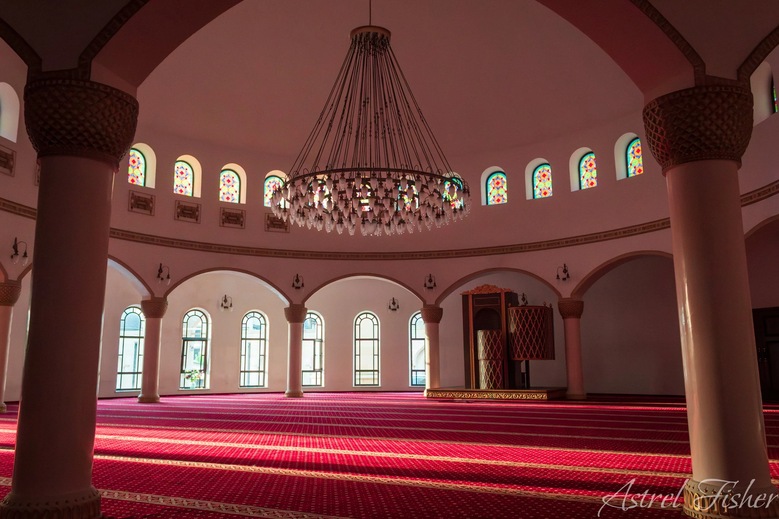 Interior of a mosque with large columns, colorful stained glass windows, and a grand chandelier hanging from the ceiling.
