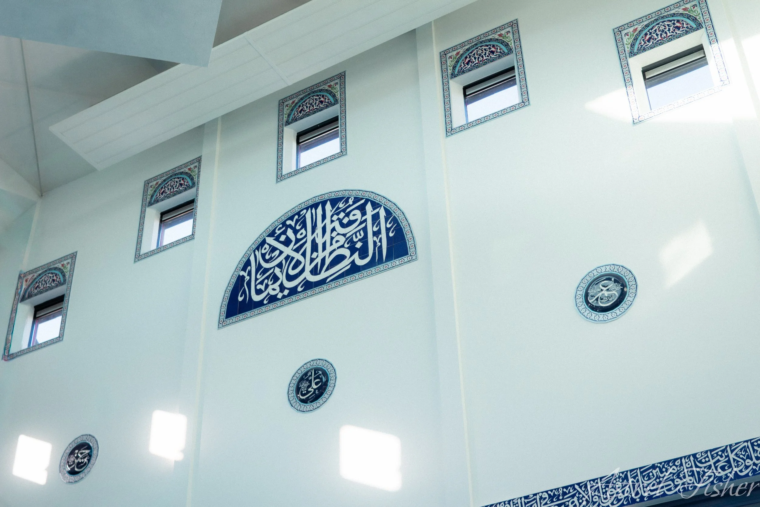 The interior of a mosque with decorative blue and white Islamic calligraphy on the walls and small rectangular windows near the ceiling.
