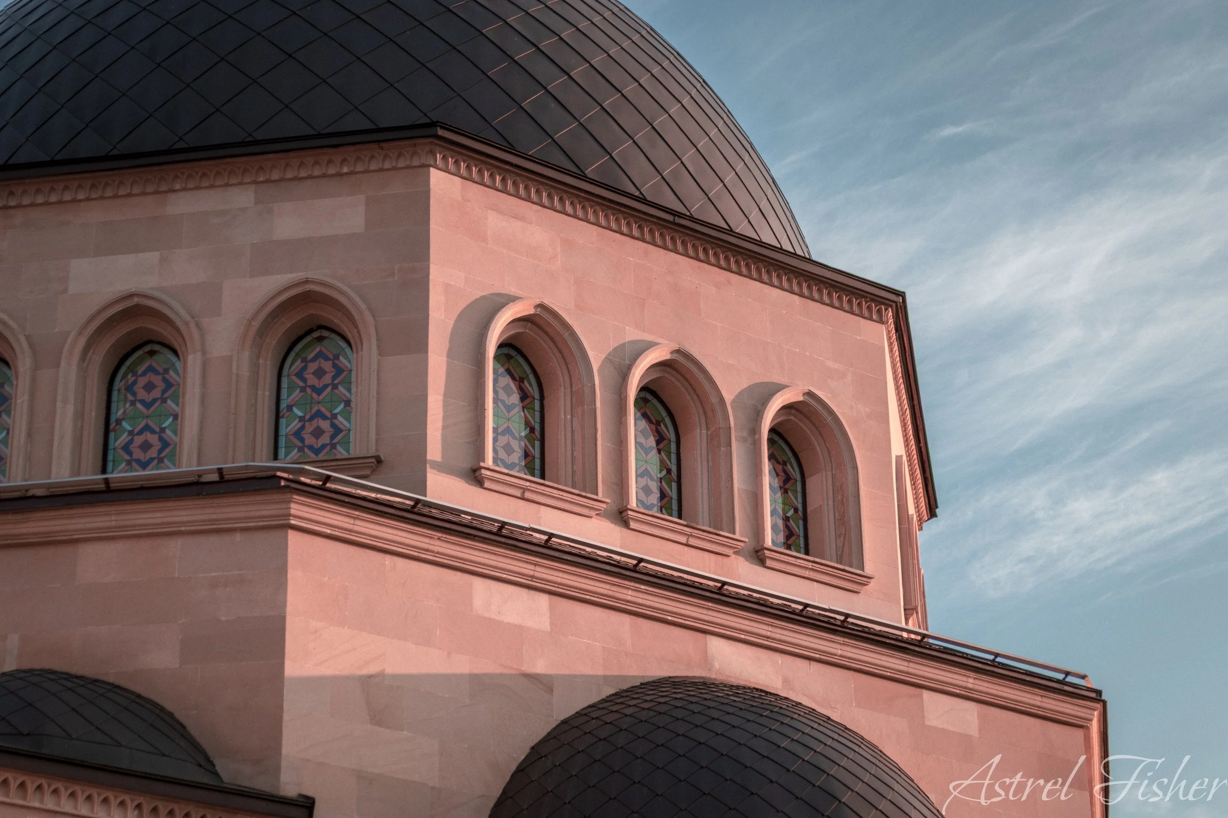 The image shows a building with a rounded dome roof and stained glass windows, set against a blue sky with some clouds.
