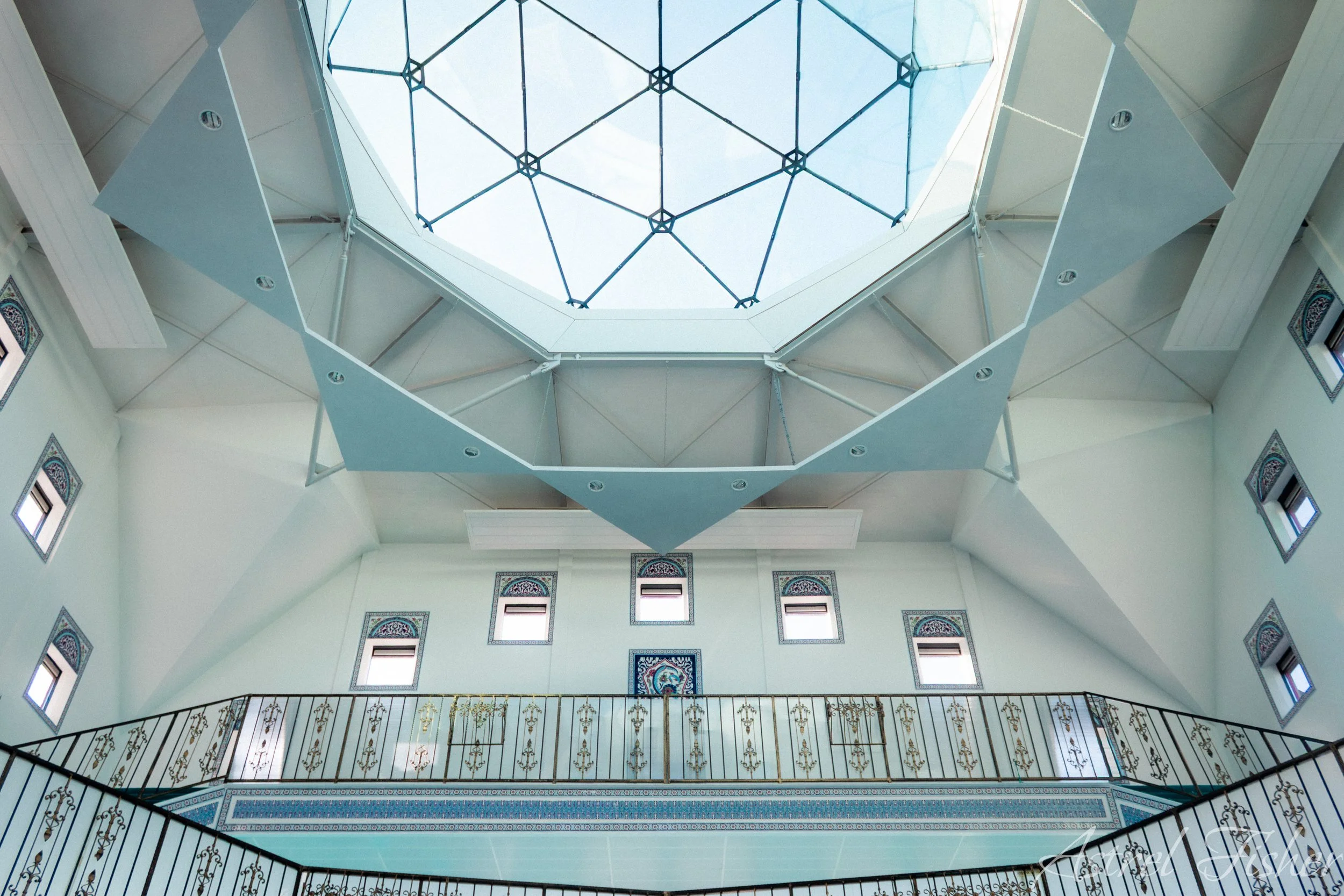 Interior view of a multi-story building with a decorative balcony, intricate railing, arched windows, and a glass ceiling with a geometric pattern.