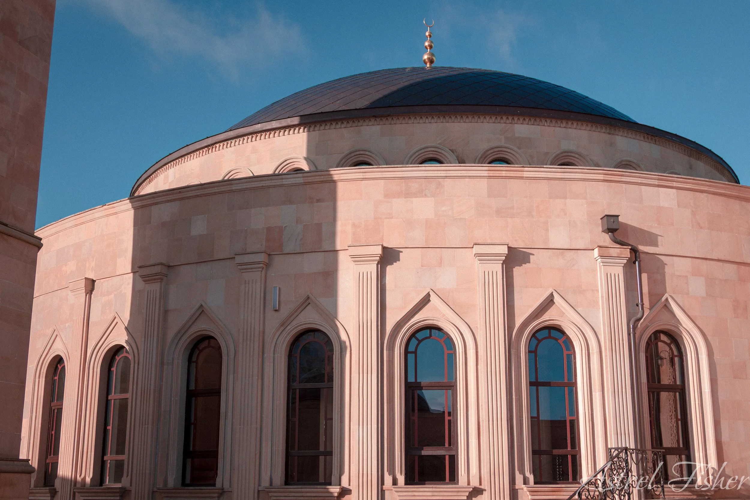 A round building with a domed roof and pink stone walls, featuring arched windows and decorative moldings, under a clear blue sky.