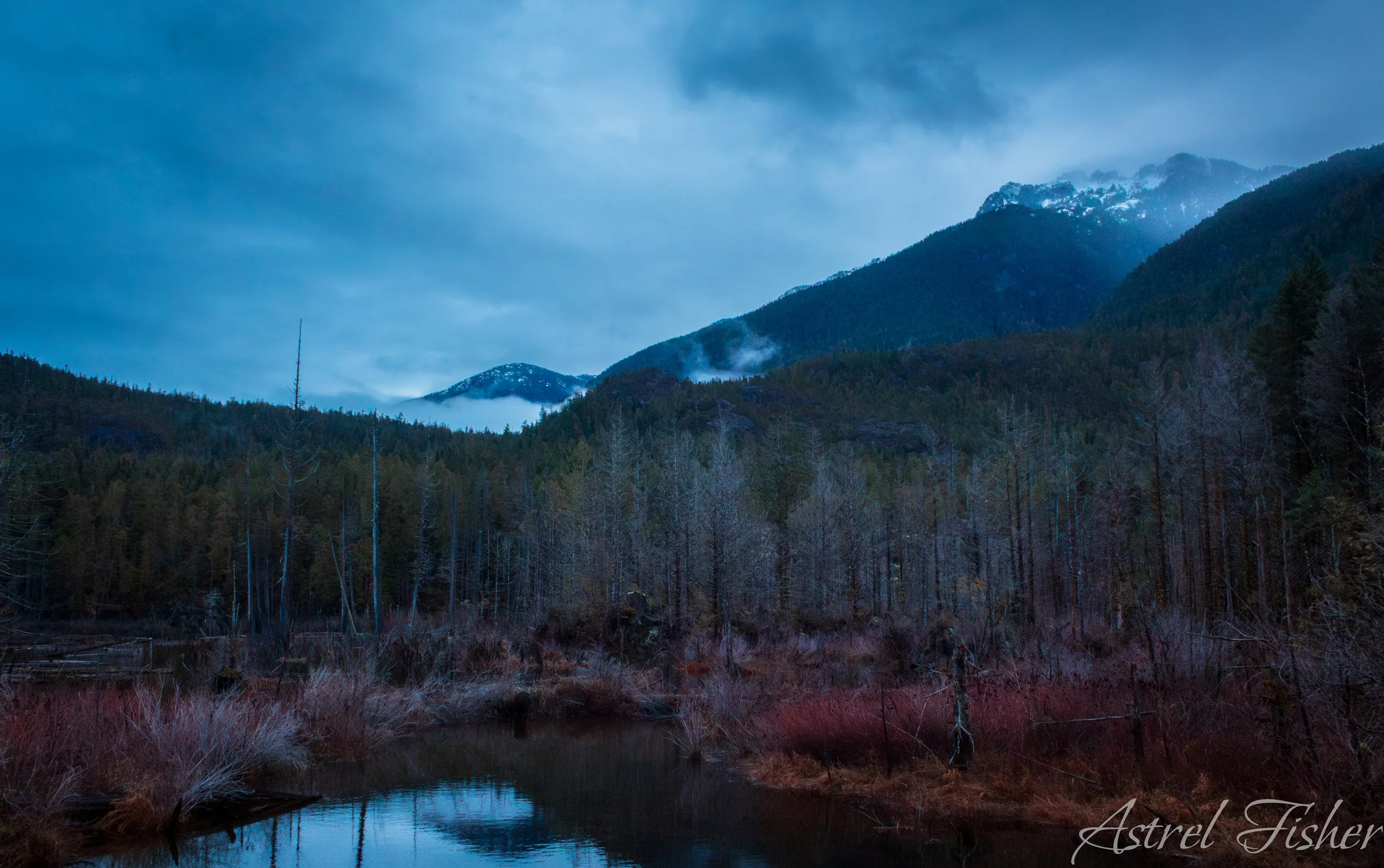 Mountain landscape with cloudy sky, fog over forested mountains, and a river in the foreground.