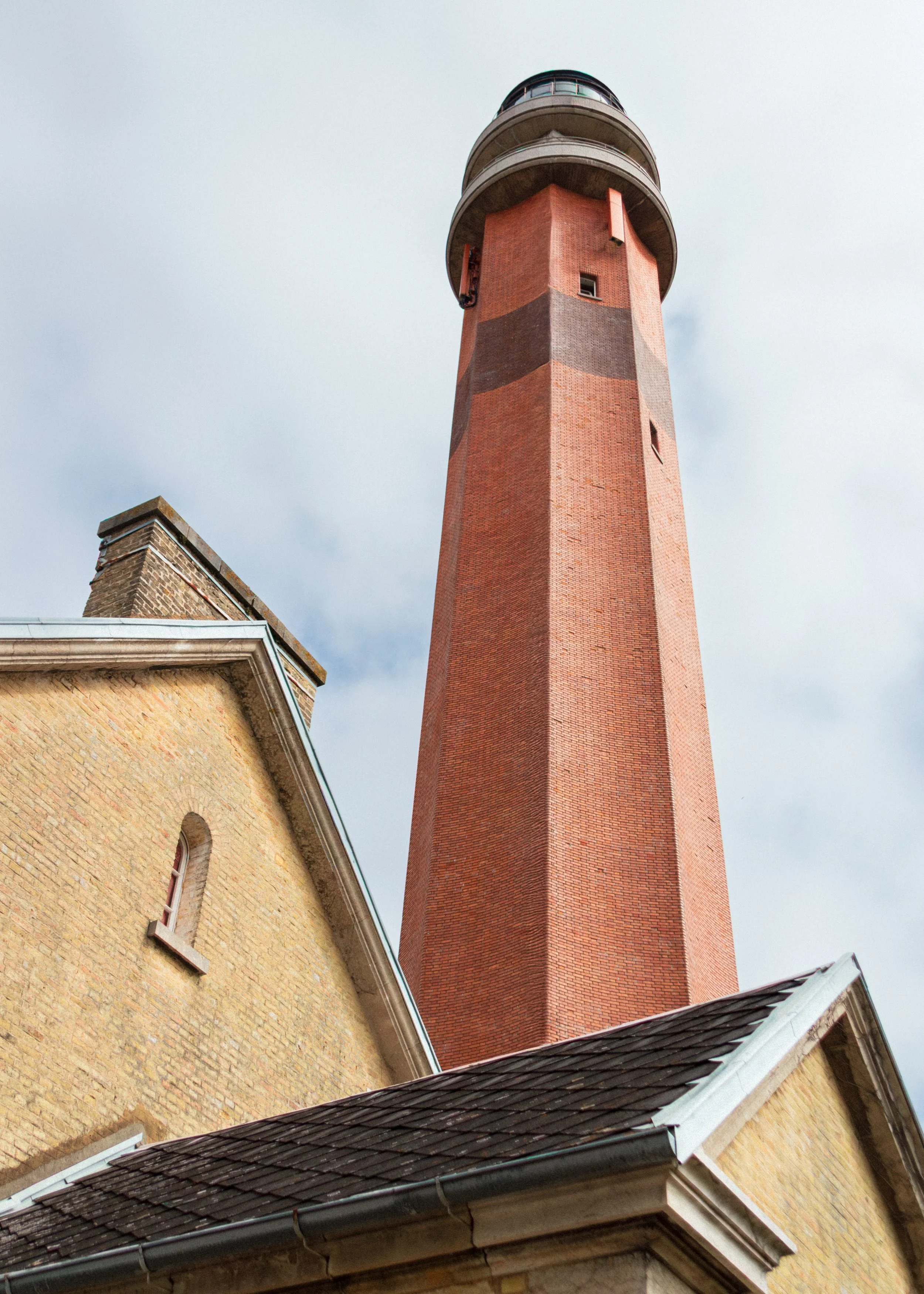 Low-angle view of a tall red brick lighthouse with a rounded top, adjacent to a yellow brick building with a sloped roof, under a cloudy sky.