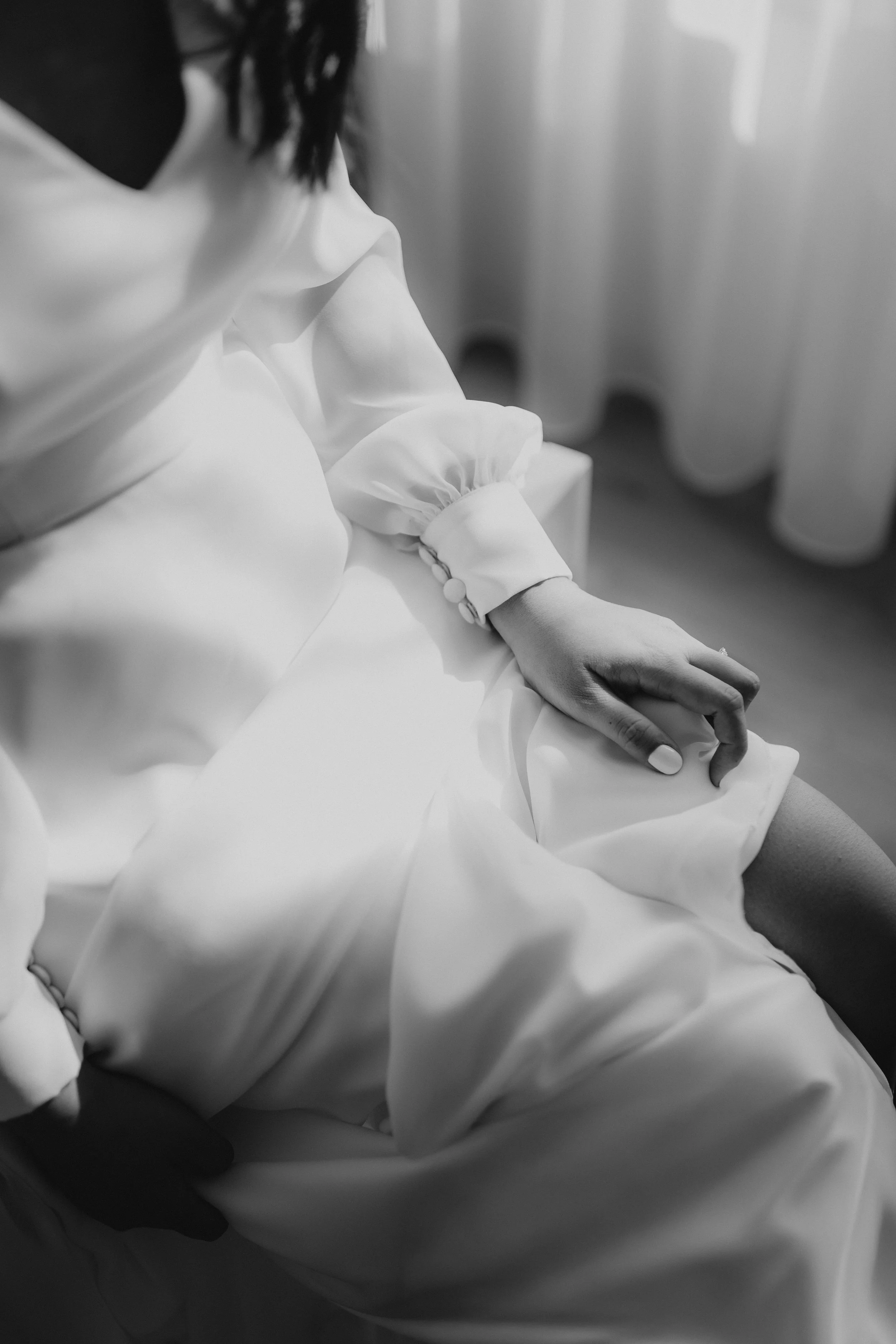 Schwarze und weiße Aufnahme einer Frau im weißen Kleid, die auf einem Stuhl sitzt, mit einer Hand auf dem Oberschenkel, vor einem Fenster mit weißen Vorhängen.