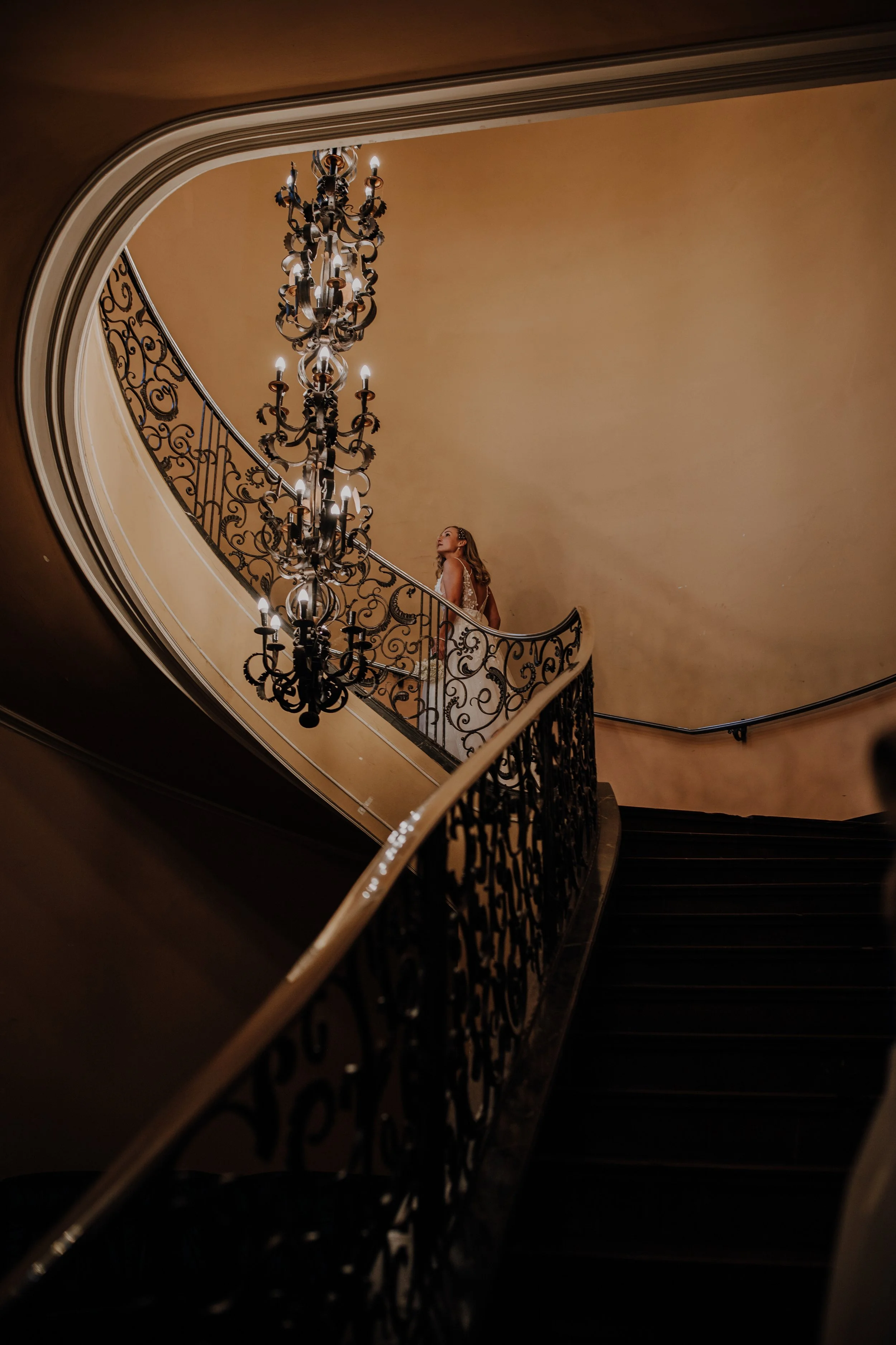 Eine elegante Treppe mit einem großen, kunstvollen Kronleuchter, eine Frau in einem weißen Kleid steht auf der Treppe, die Wand im Hintergrund ist beige.