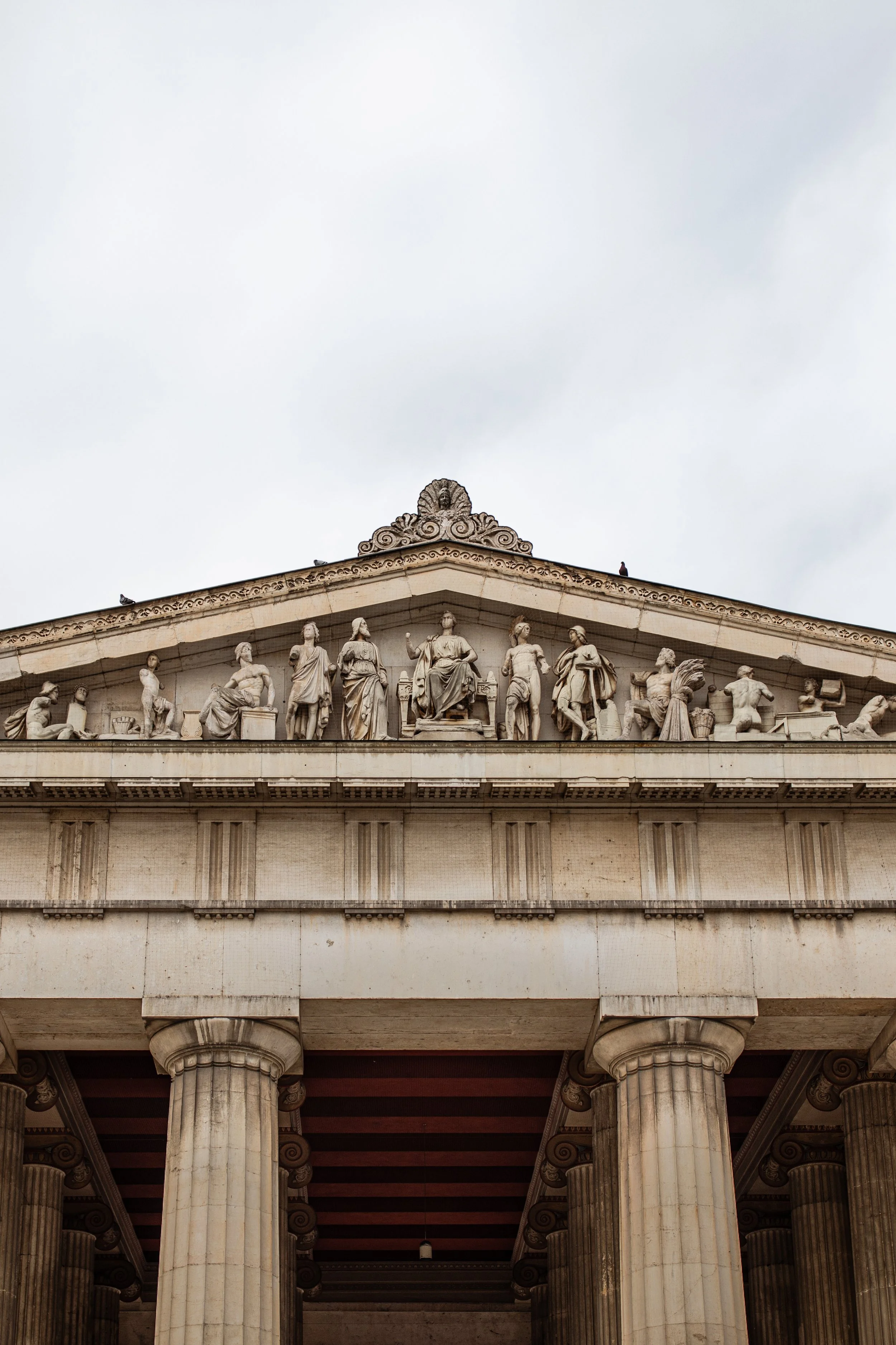 Das Bild zeigt die Fratentreppe des Brandenburger Tors in Berlin, mit einer Reihe von steinernen Skulpturen auf dem oberen Giebel, außerdem einige Vögel auf dem Dach.