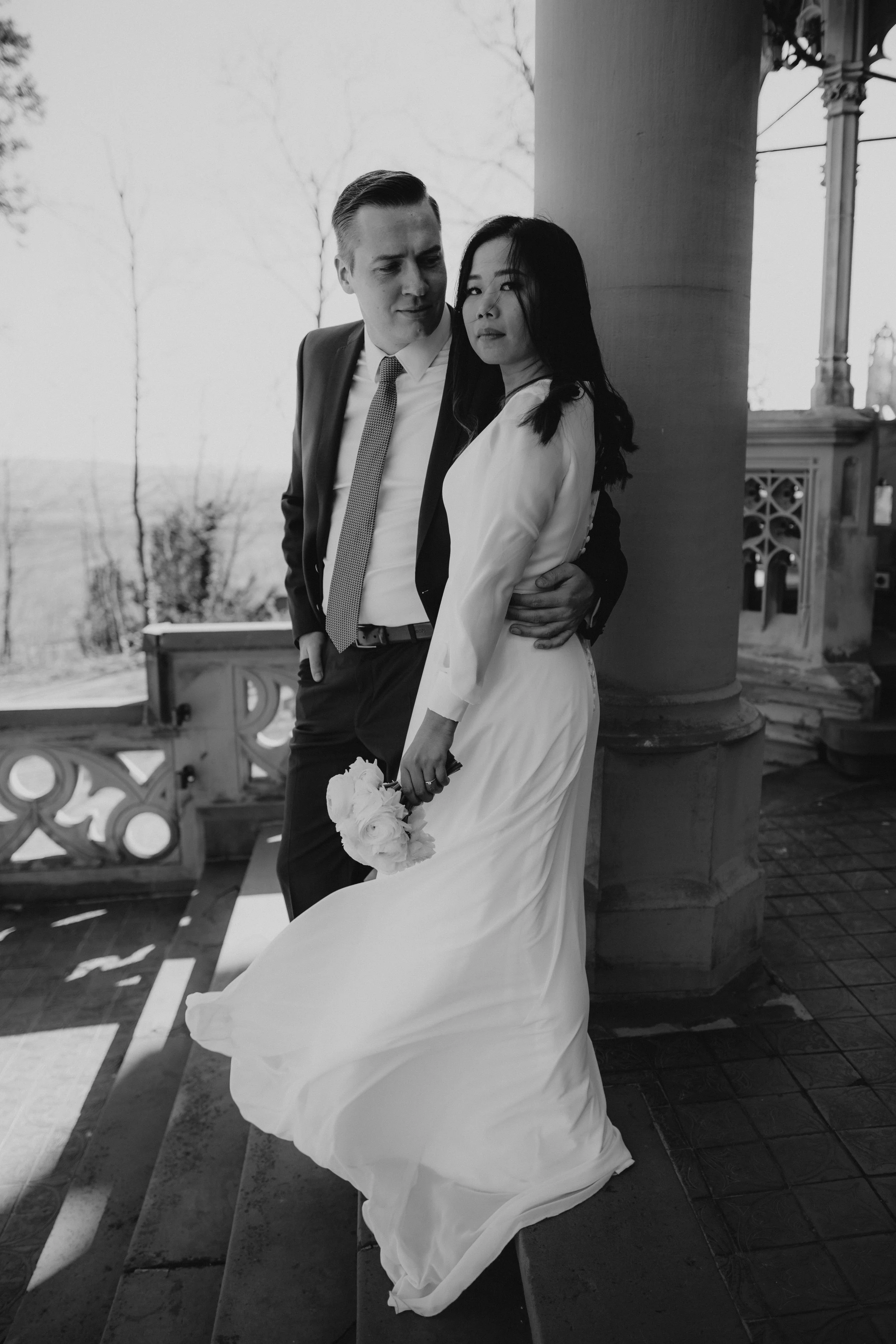 Ein schwarz-weiß-Foto eines Paares in formeller Kleidung, die auf einer Veranda mit Säulen stehen. Die Frau trägt ein langes weißes Kleid und hält einen Blumenstrauß, während der Mann einen dunklen Anzug trägt und sie umarmt.