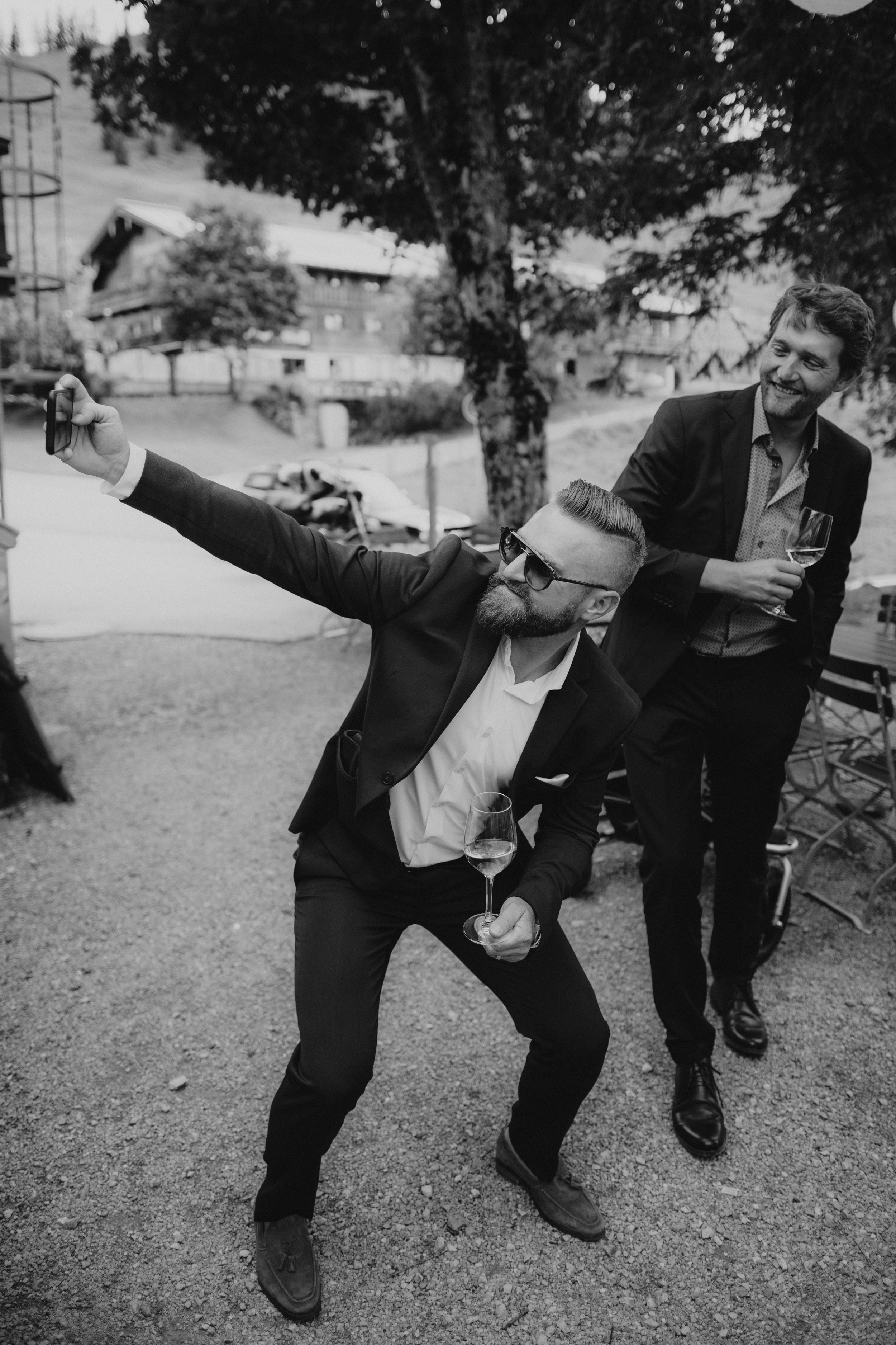 Zwei Männer in Anzügen, einer macht ein Selfie, der andere lacht und hält ein Weinglas, bei einem Treffen im Freien, im Hintergrund Bäume, Häuser und ein Auto, Schwarz-Weiß-Foto.