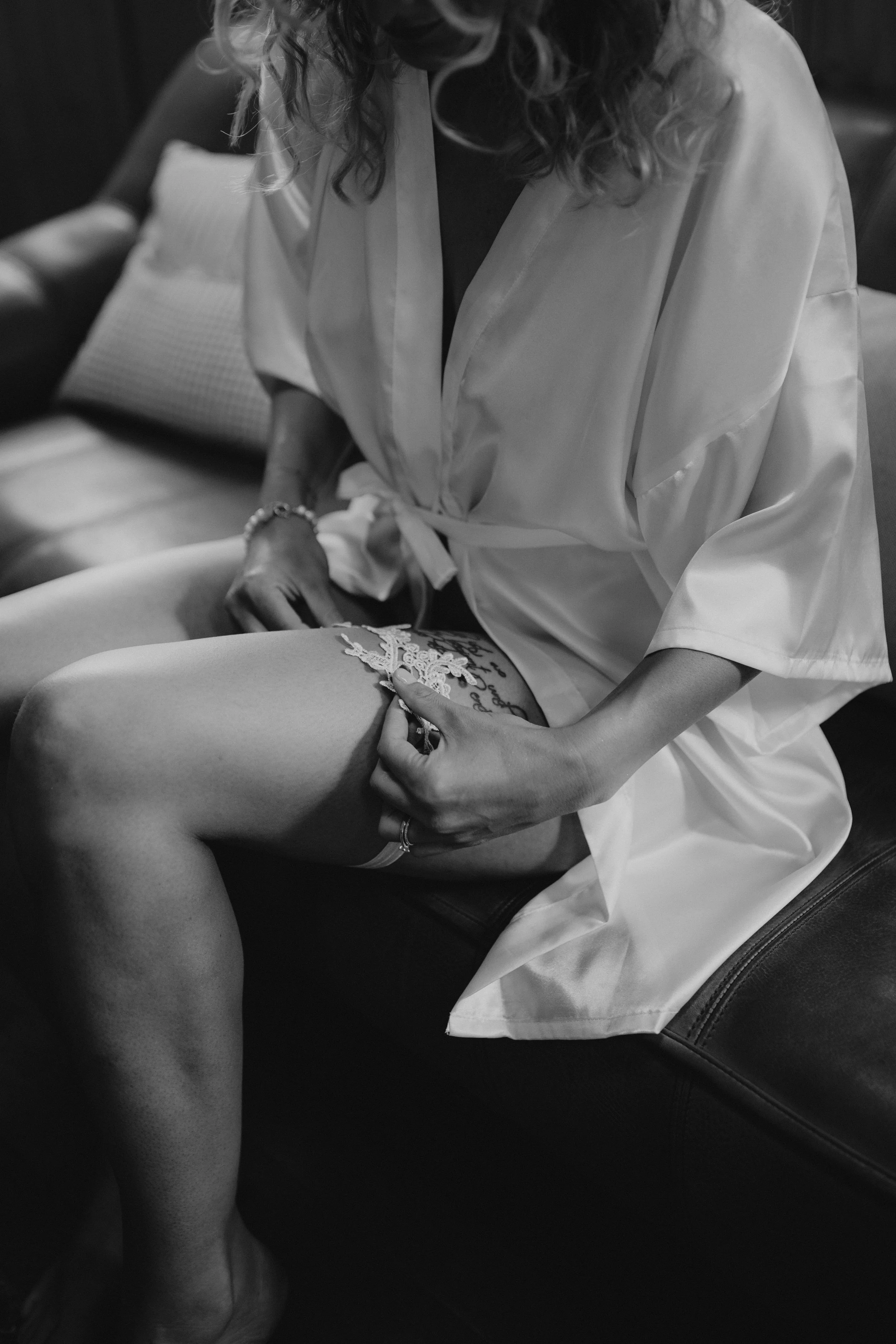 Schwarz-Weiß-Foto einer Frau in einem weißen Bademantel, die ein Tattoo auf ihrem Oberschenkel zeigt, während sie auf einem schwarzen Sitz sitzt.