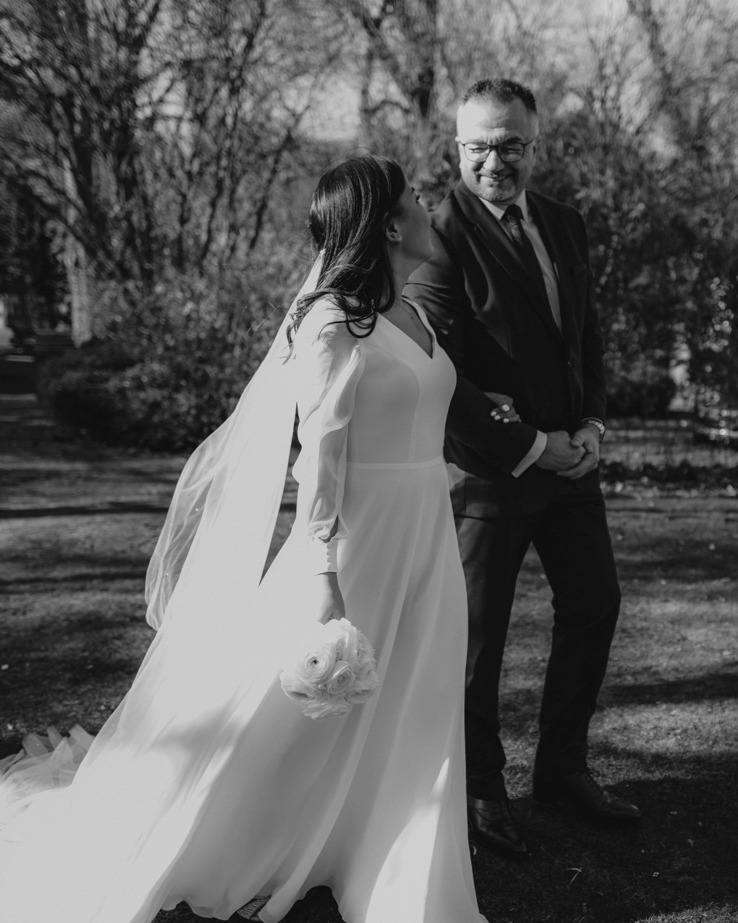 Ein Brautpaar im Freien, die Braut hält einen Blumenstrauß, der Bräutigam trägt einen dunklen Anzug und eine Krawatte.