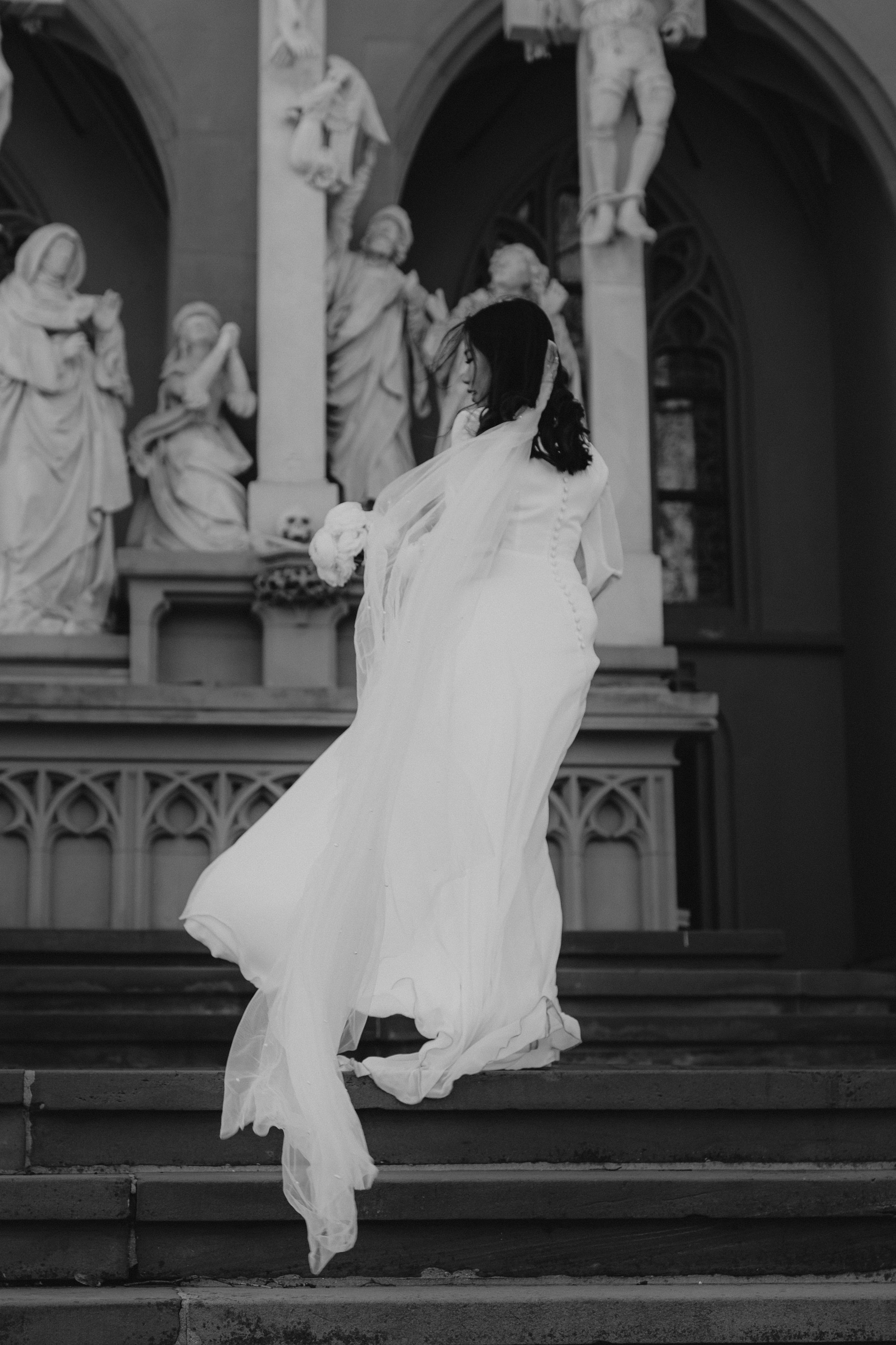 Schwarz-weiß-Foto einer Frau in einem langen weißen Kleid, die die Treppe eines historischen Kircheninneren hinaufgeht, umgeben von religiösen Skulpturen und Figuren.
