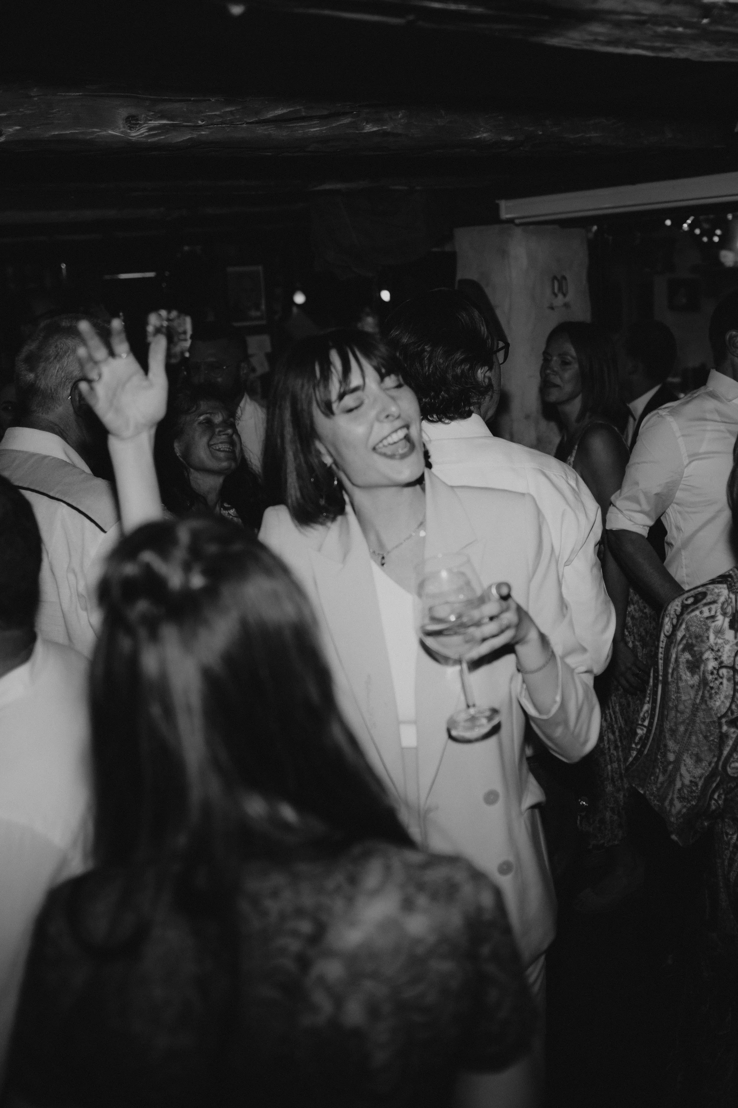 Menschen feiern in einer Bar, lachen und tanzen, eine Frau im Vordergrund hält ein Glas Wein.