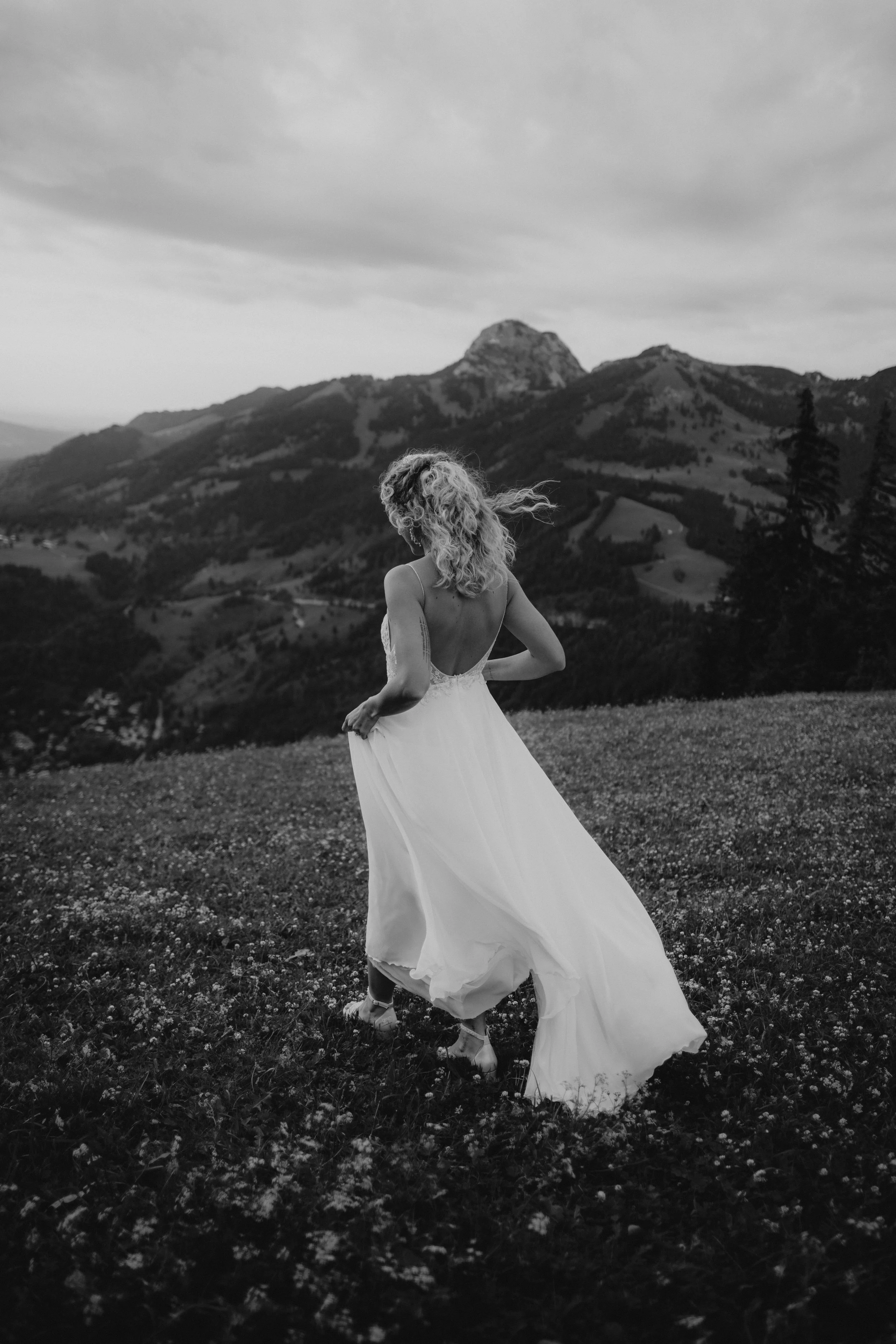 Eine Frau mit einer langen weißen Hochzeitstracht steht auf einer Blumenwiese mit Bergpanorama im Hintergrund, im schwarzen und weißen Foto.