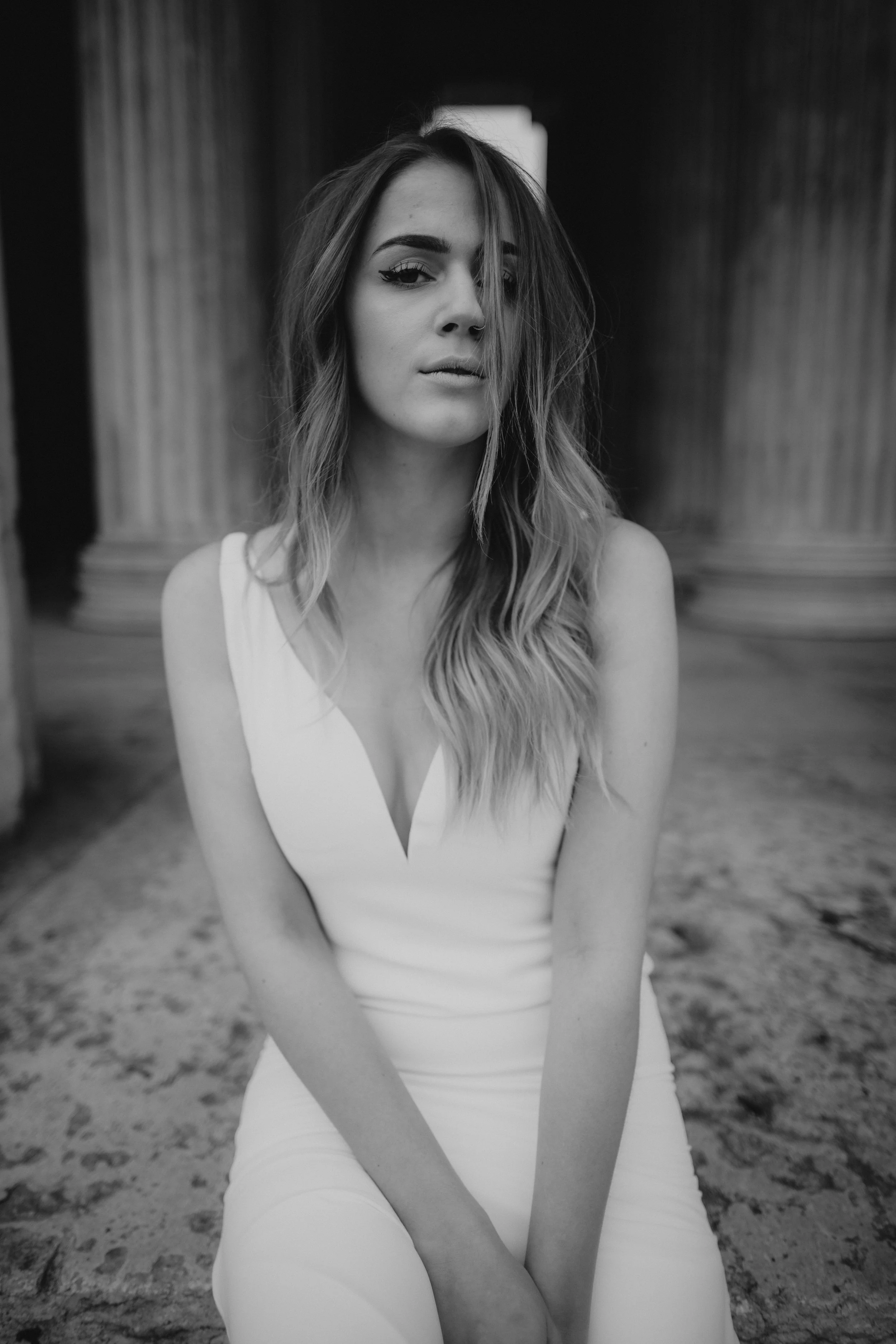 Schwarz-weiß-Foto einer jungen Frau in einem weißen Kleid, sitzend vor antiken Säulen, mit langem, gewelltem Haar und Blick zur Seite.