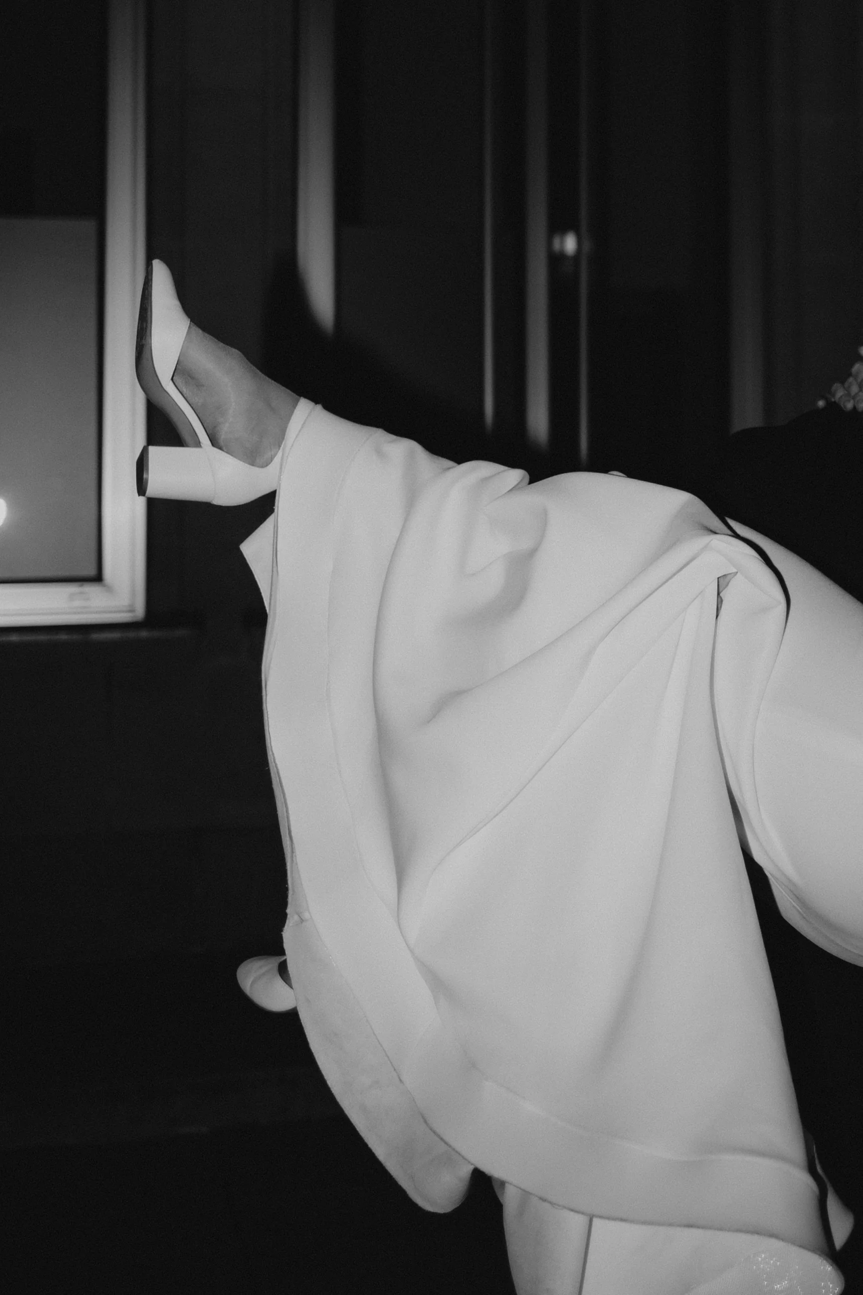 Schwarz-weiß-Foto einer Person, die eine weiße Jacke und Schuhe trägt, mit einem Bein in die Luft gestreckt, vor einem dunklen Hintergrund.