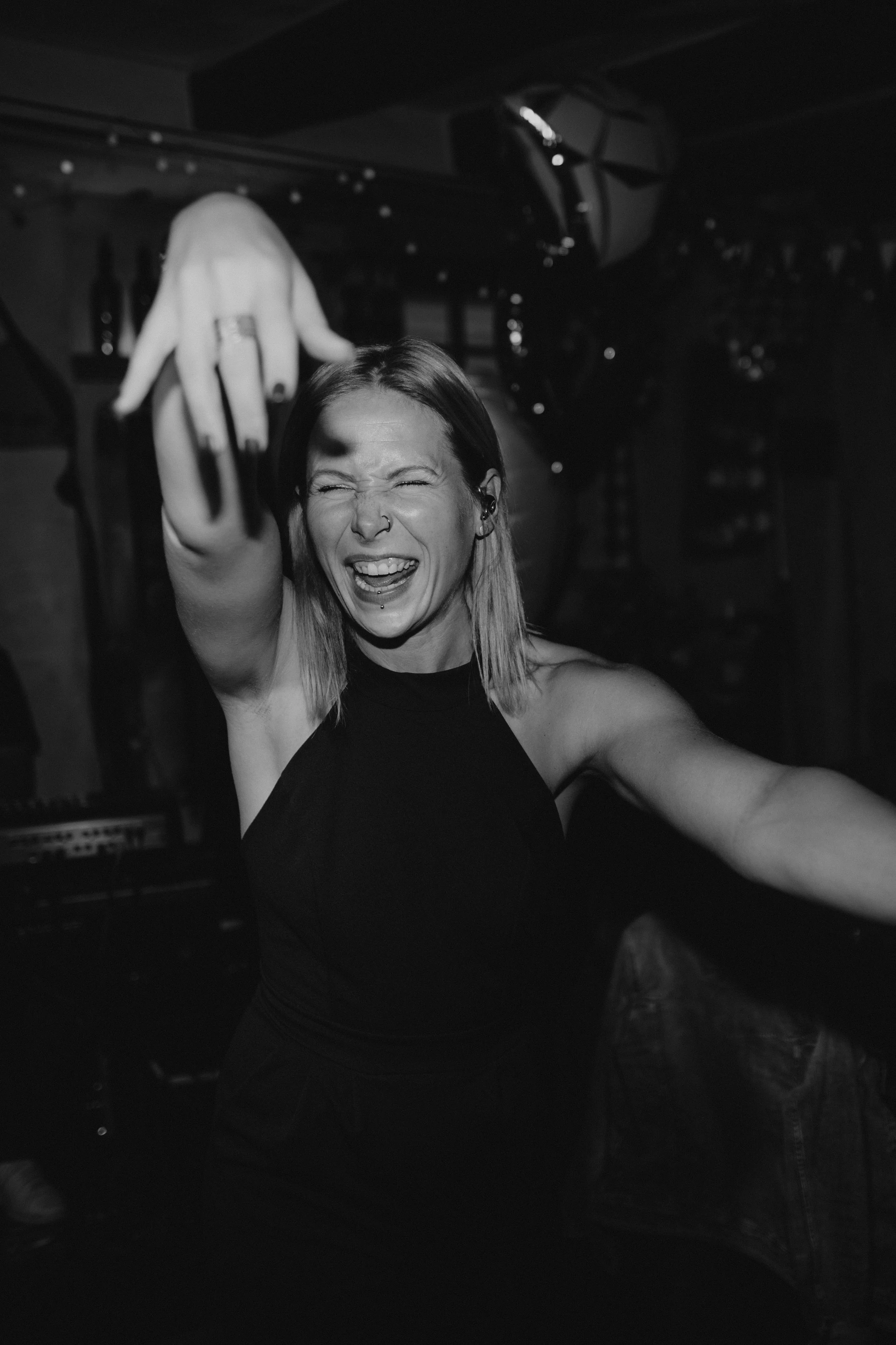 Eine lachende junge Frau mit Piercings und dunklem Oberteil, die in einer dunklen Bar oder Club tanzt oder schaukelt.