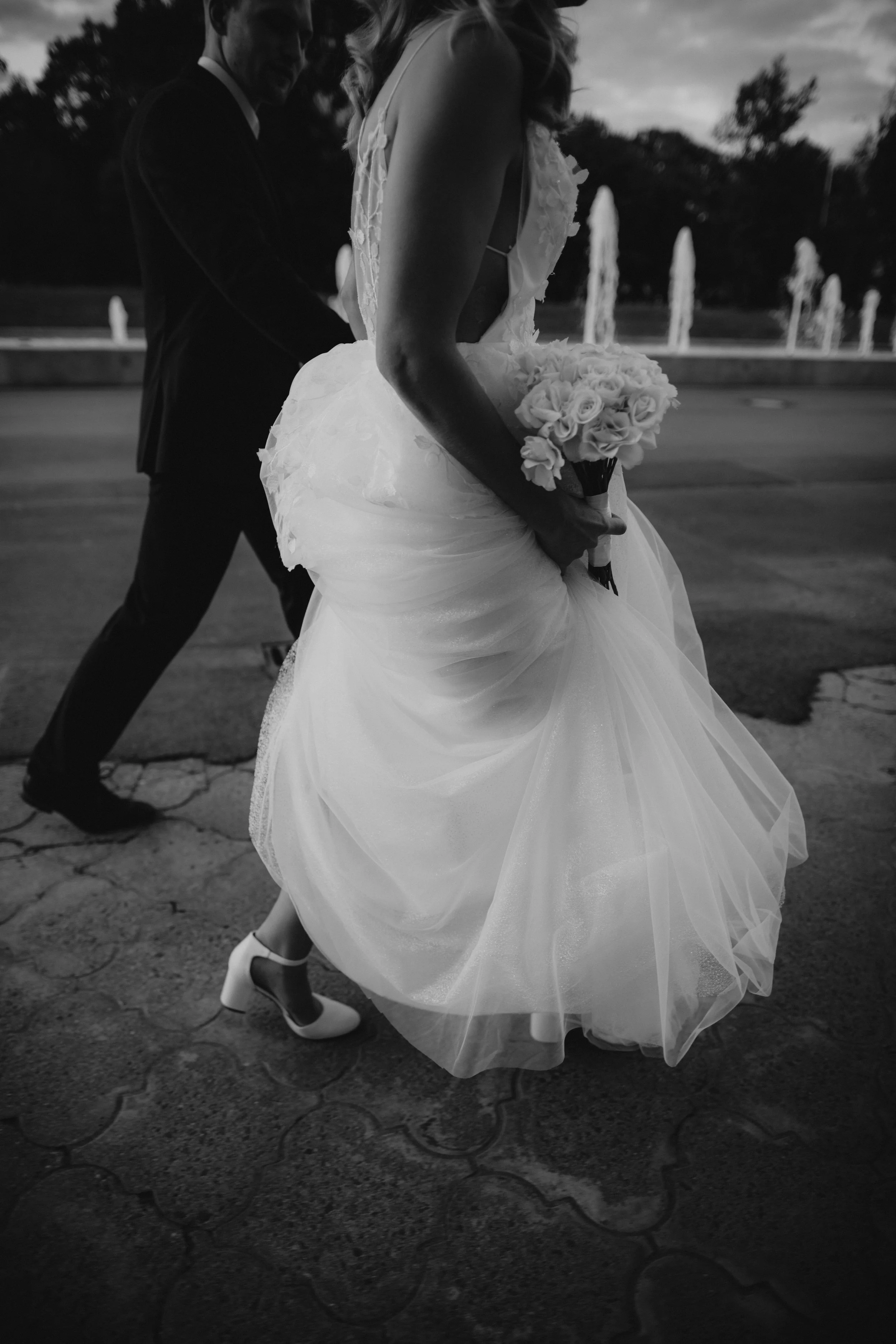 Braut in Hochzeitskleid mit Blumenstrauß, begleitet von Bräutigam, bei Sonnenuntergang vor Springbrunnen.