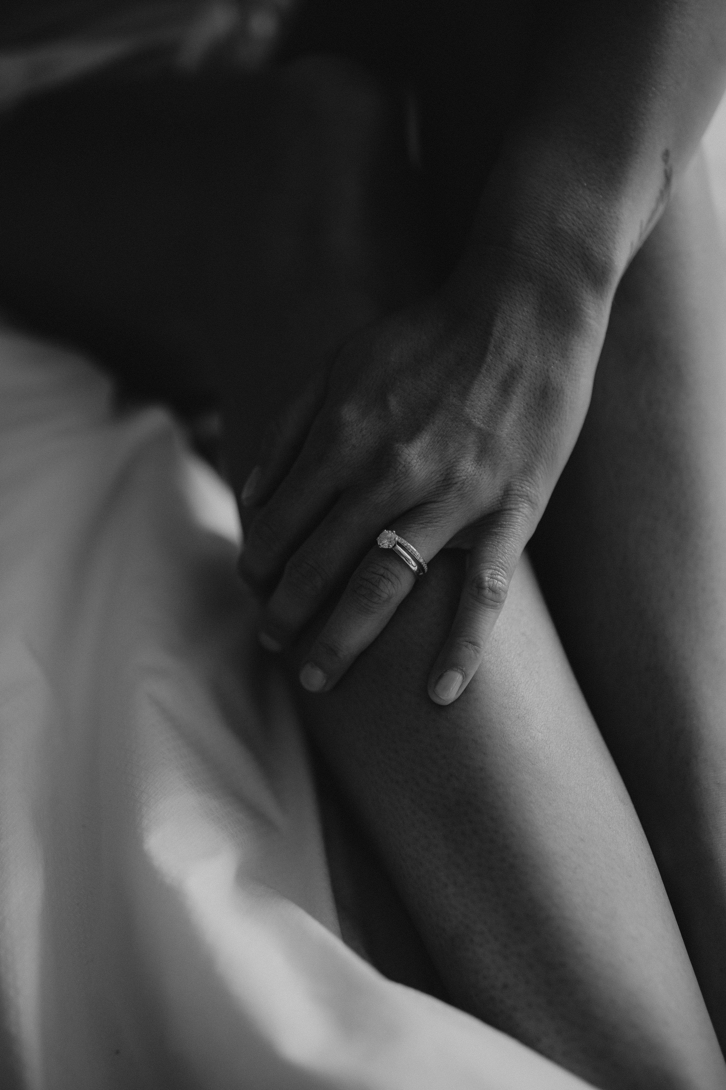 Schwarz-weiß-Foto einer Hand mit einem Verlobungsring, der auf das Bein einer Person gelegt ist.