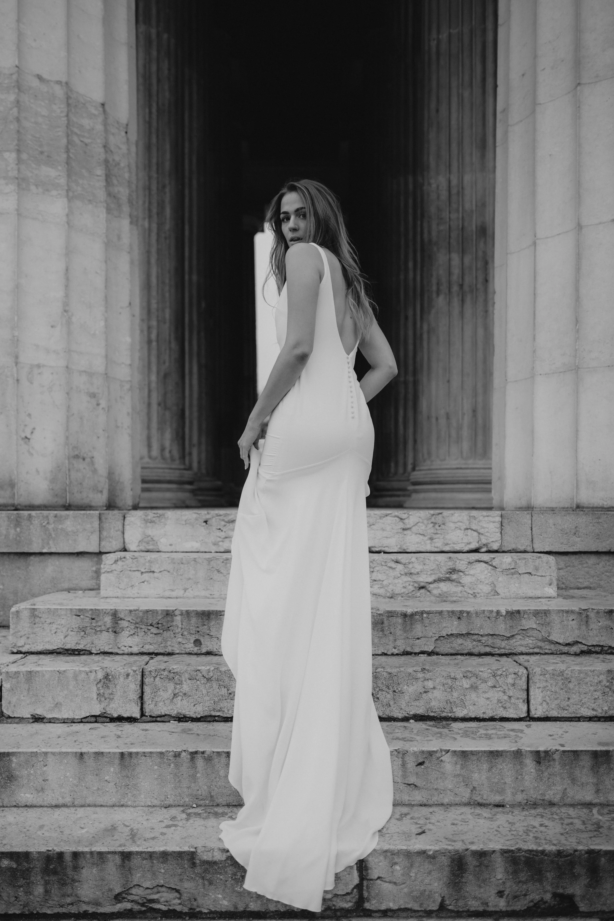 Schwarz-weiß-Foto einer jungen Frau in einem langen weißen Kleid, die auf den Stufen vor einem antiken Gebäude steht.