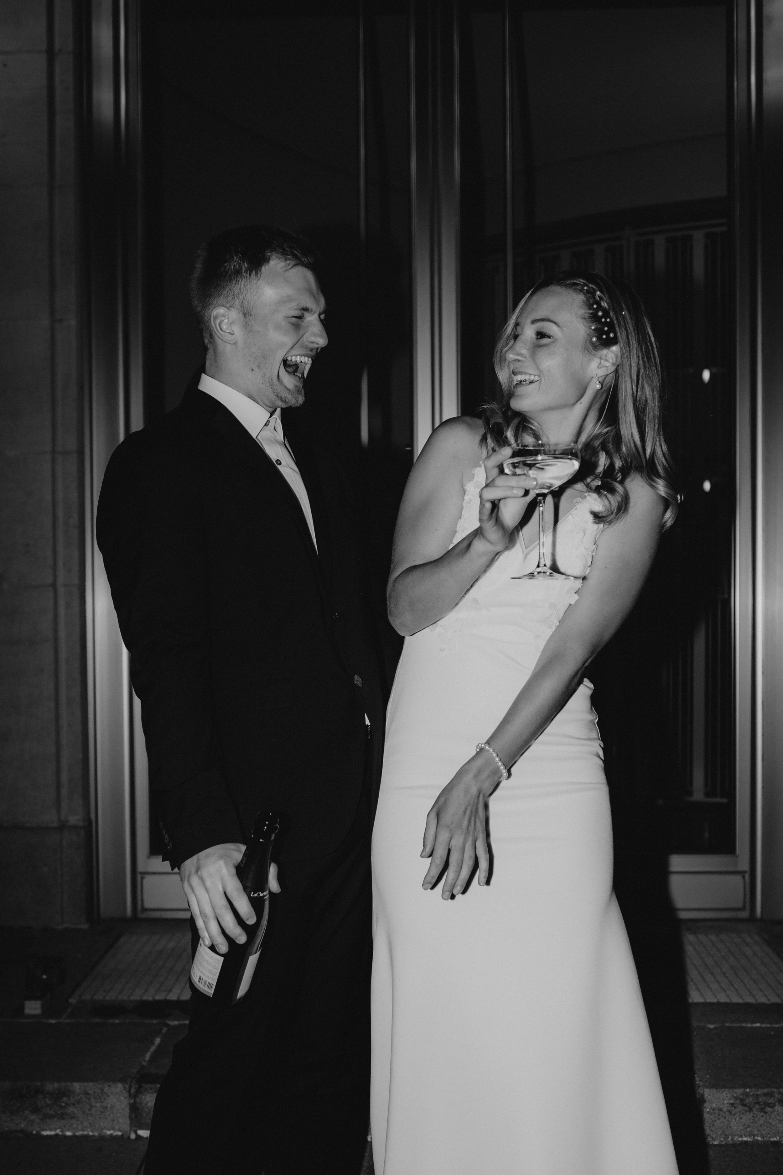 Ein lachendes Paar in formeller Kleidung bei einer Feier, wahrscheinlich einer Hochzeit, vor einem Gebäude, beide halten Gläser mit Champagner.