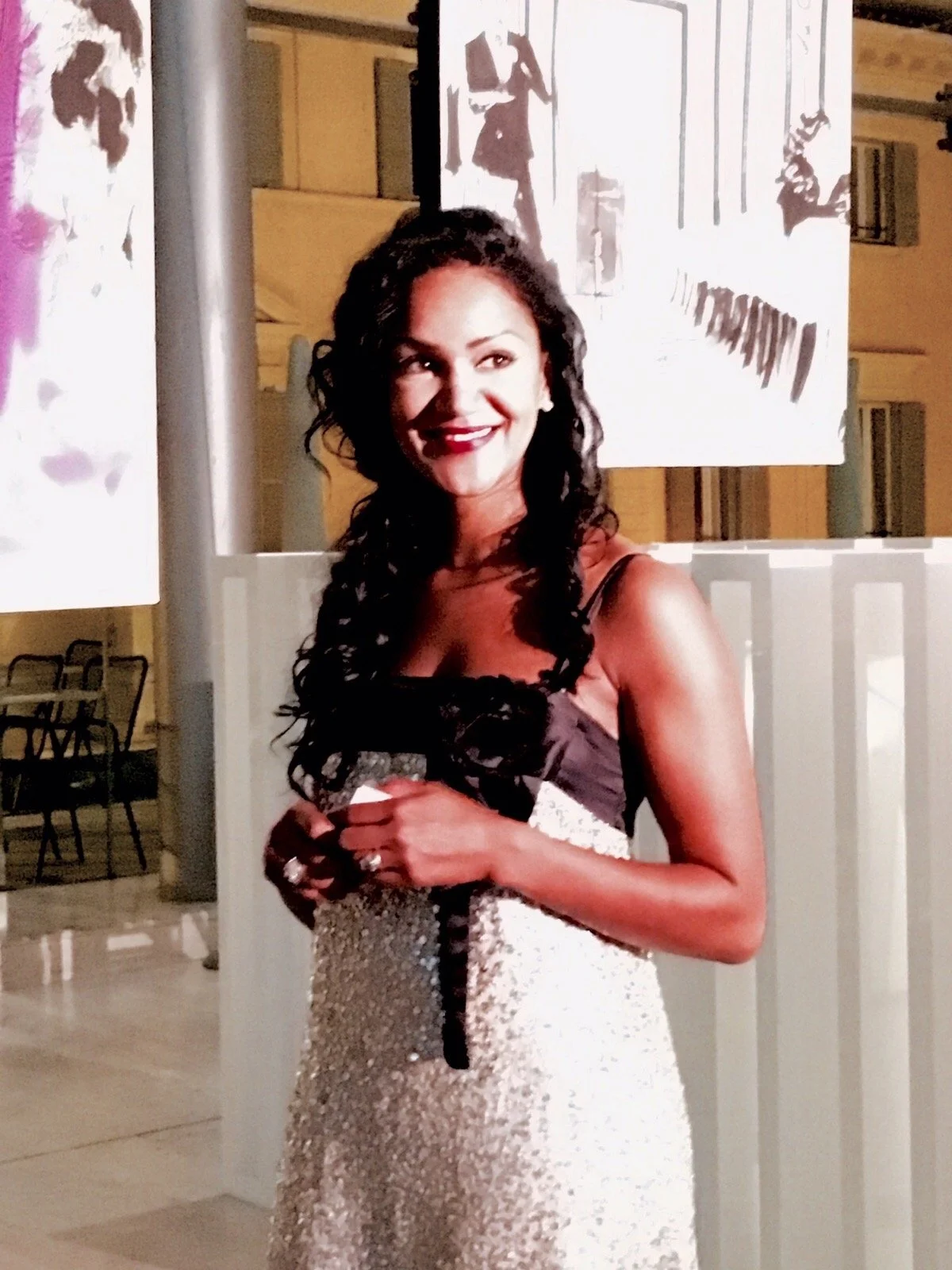 A woman with long dark curly hair smiling, in front of her paintings, wearing a black and white dress with black lace straps.
