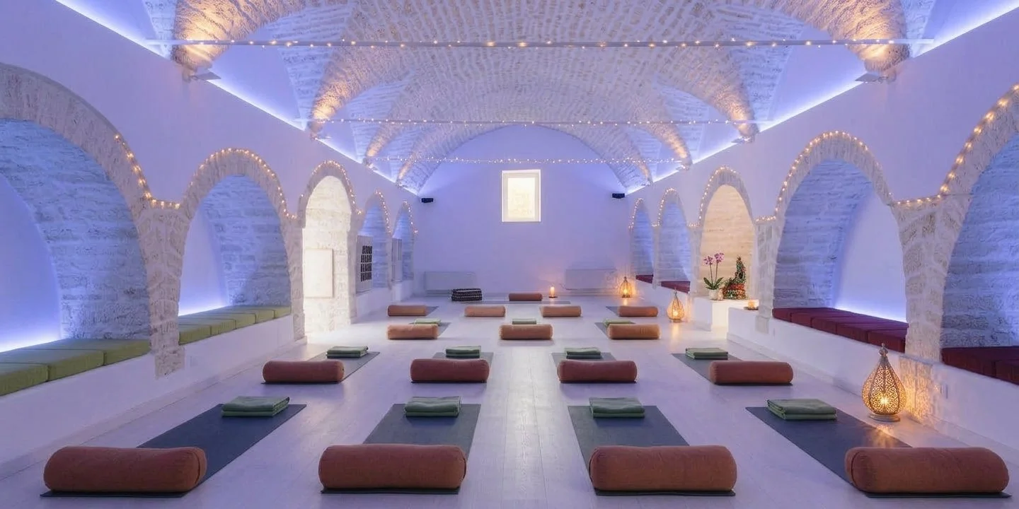 A serene, well-lit yoga studio with white stone walls, arched alcoves, and soft ambient lighting. Yoga mats and bolsters are arranged on the floor with folded towels, and decorative lanterns are placed along the sides.